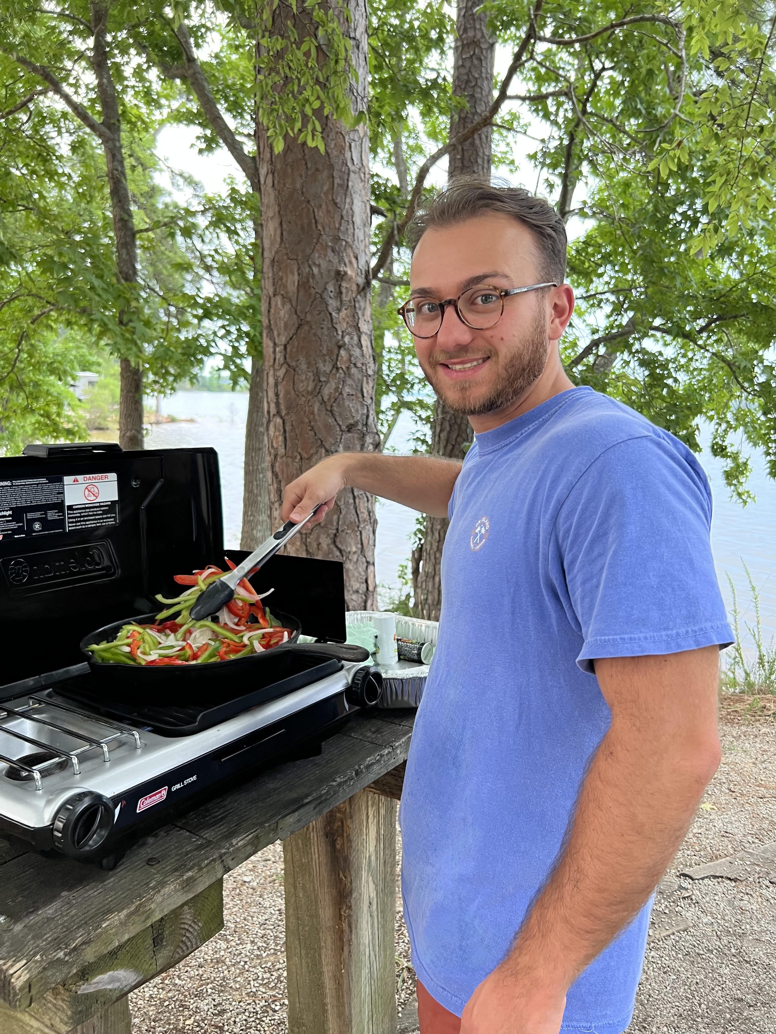 A man wearing glasses and a blue t-shirt is grilling vegetables on a portable gas grill outdoors near a lake, with trees in the background.