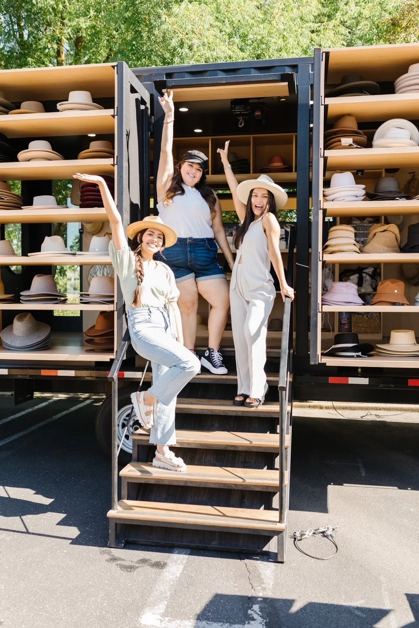 Three women pose happily on steps in front of a hat display truck, wearing summer clothes and hats, with green trees in the background.