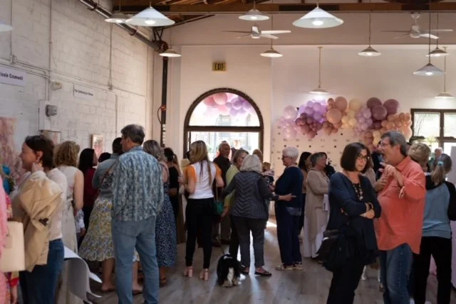 People gathered in a spacious room with high ceilings, white walls, and large windows, decorated with pink and purple balloon arrangements, likely for a celebration or event.