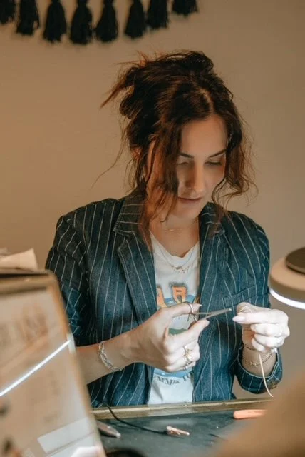 Woman knitting at a table, with a black and white striped blazer, in a room with beige walls and a black decorative item hanging on the wall.