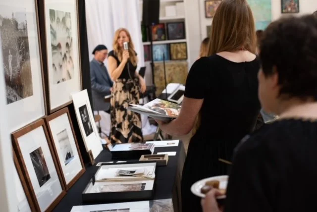 People at an art gallery viewing framed photographs and artwork on display, with a woman taking a photo in the background.