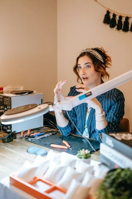 Woman with curly hair wearing a headband and striped blazer, speaking with hand gestures, sitting at a cluttered desk with electronics and tools, in a room with beige walls and black tassel wall decor.
