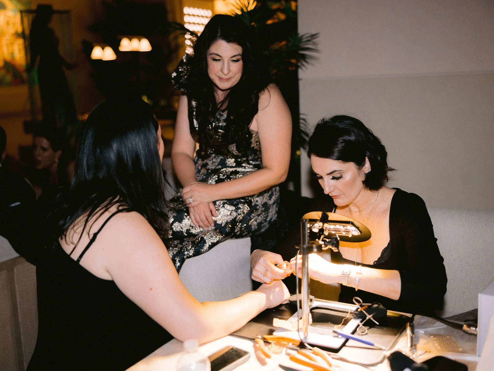 Women sitting at a table while one woman gets a manicure from a manicurist using a nail lamp, with others watching.