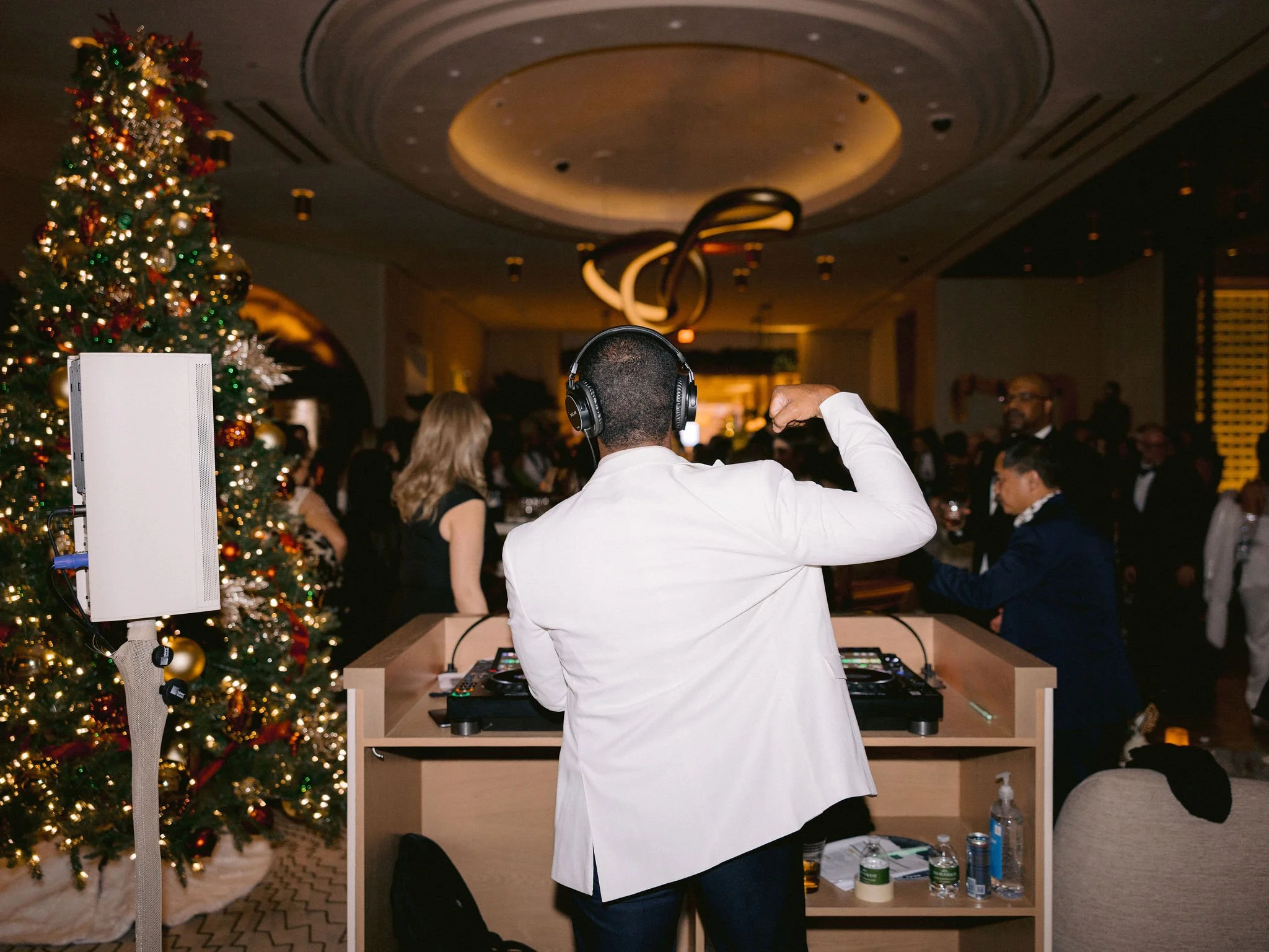 A DJ wearing a white shirt and headphones is playing music at a holiday party, with a decorated Christmas tree and guests dancing in the background.