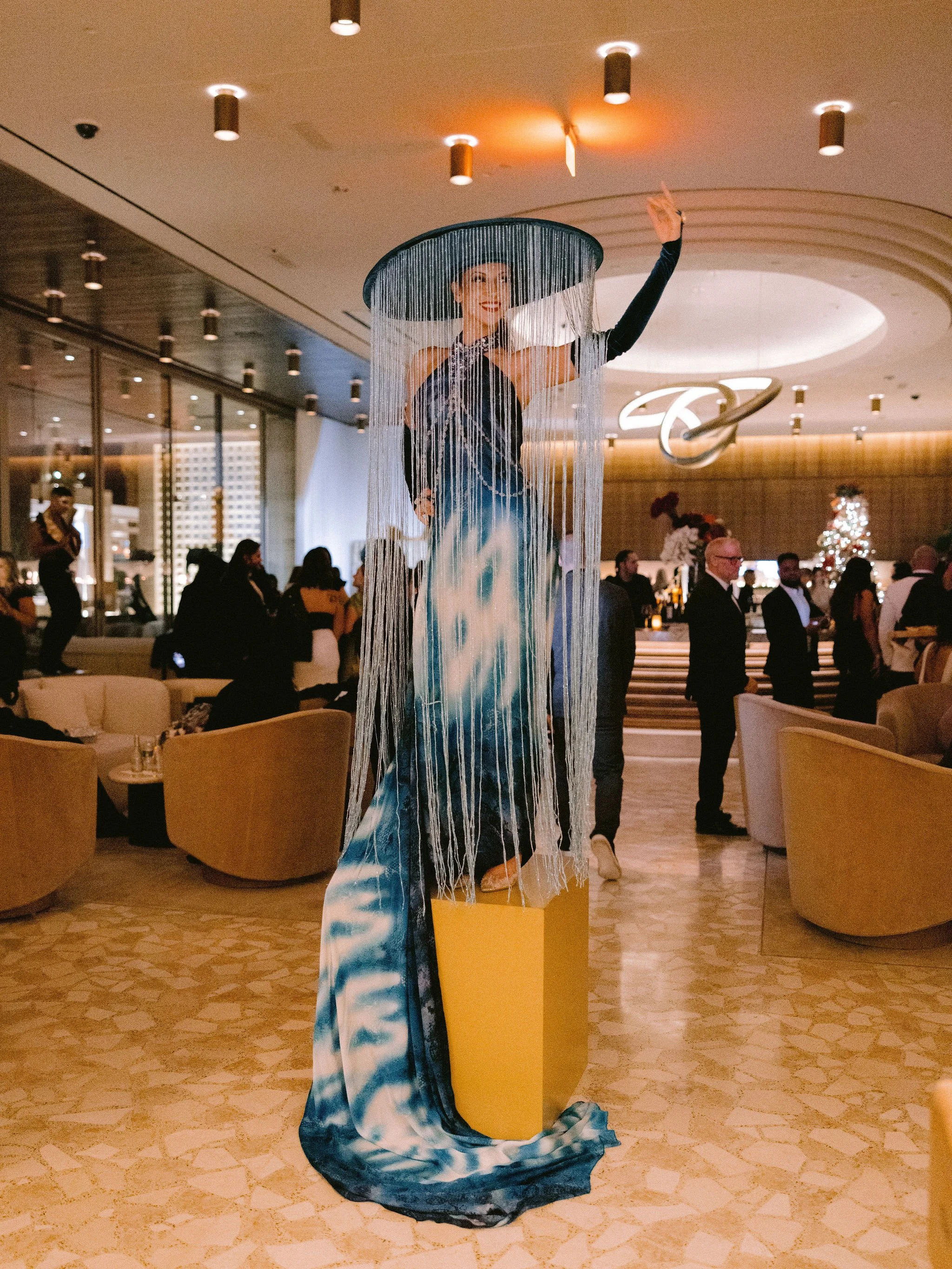 A performer in a dramatic outfit with a large hat and flowing dress, standing on a yellow pedestal inside a decorated venue with guests in formal attire.