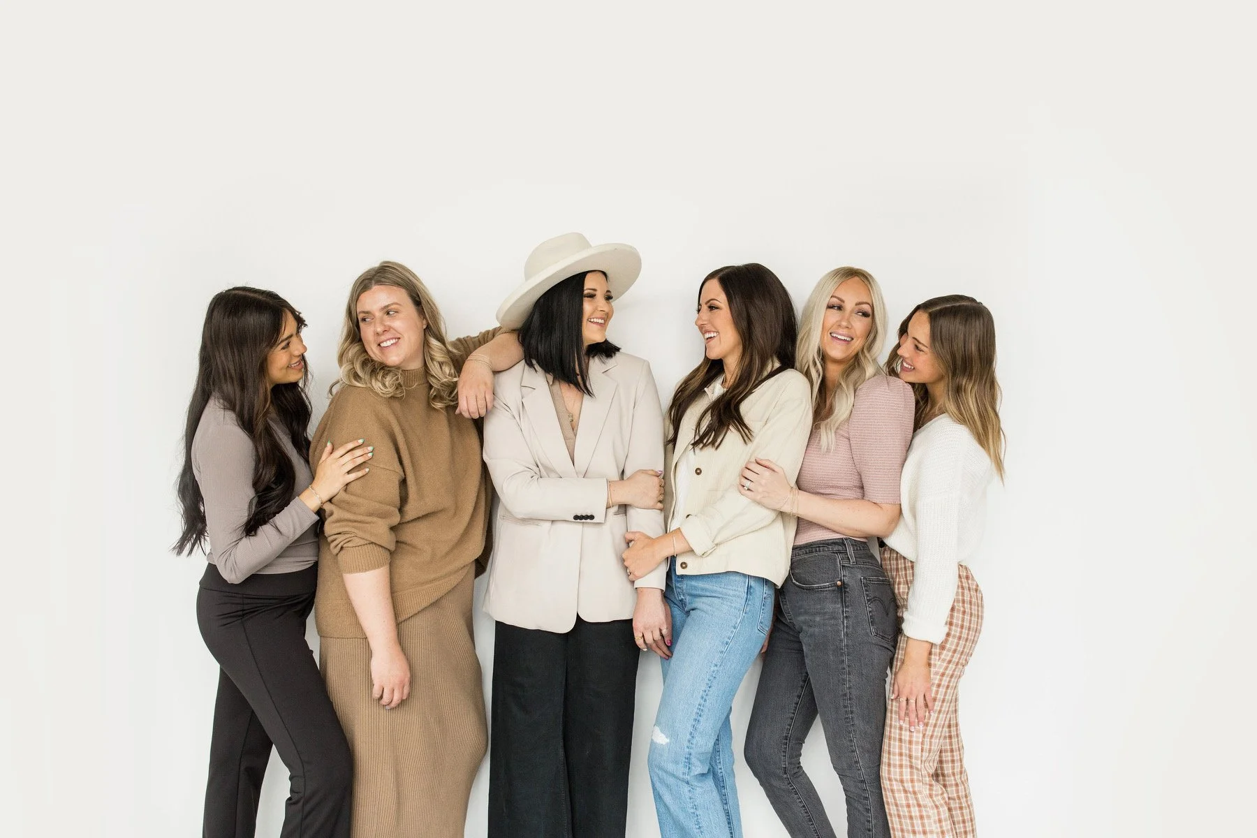 A group of six women standing together against a plain white wall, smiling and interacting with each other. They are dressed in casual and semi-formal attire.