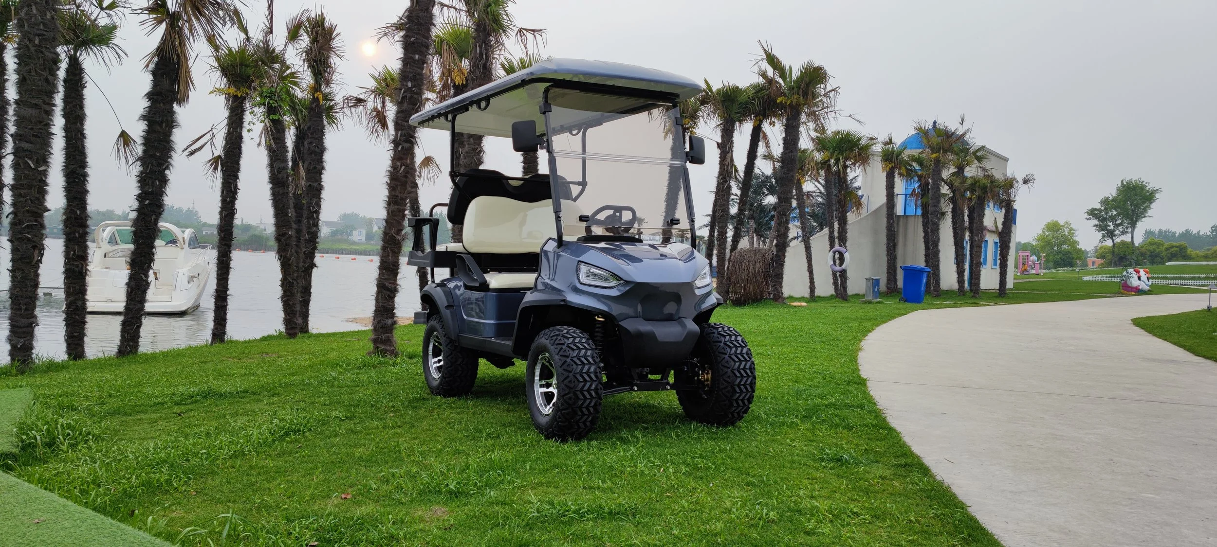 Golf cart parked on a grassy area near a pathway with palm trees in the background and a lake with a boat.