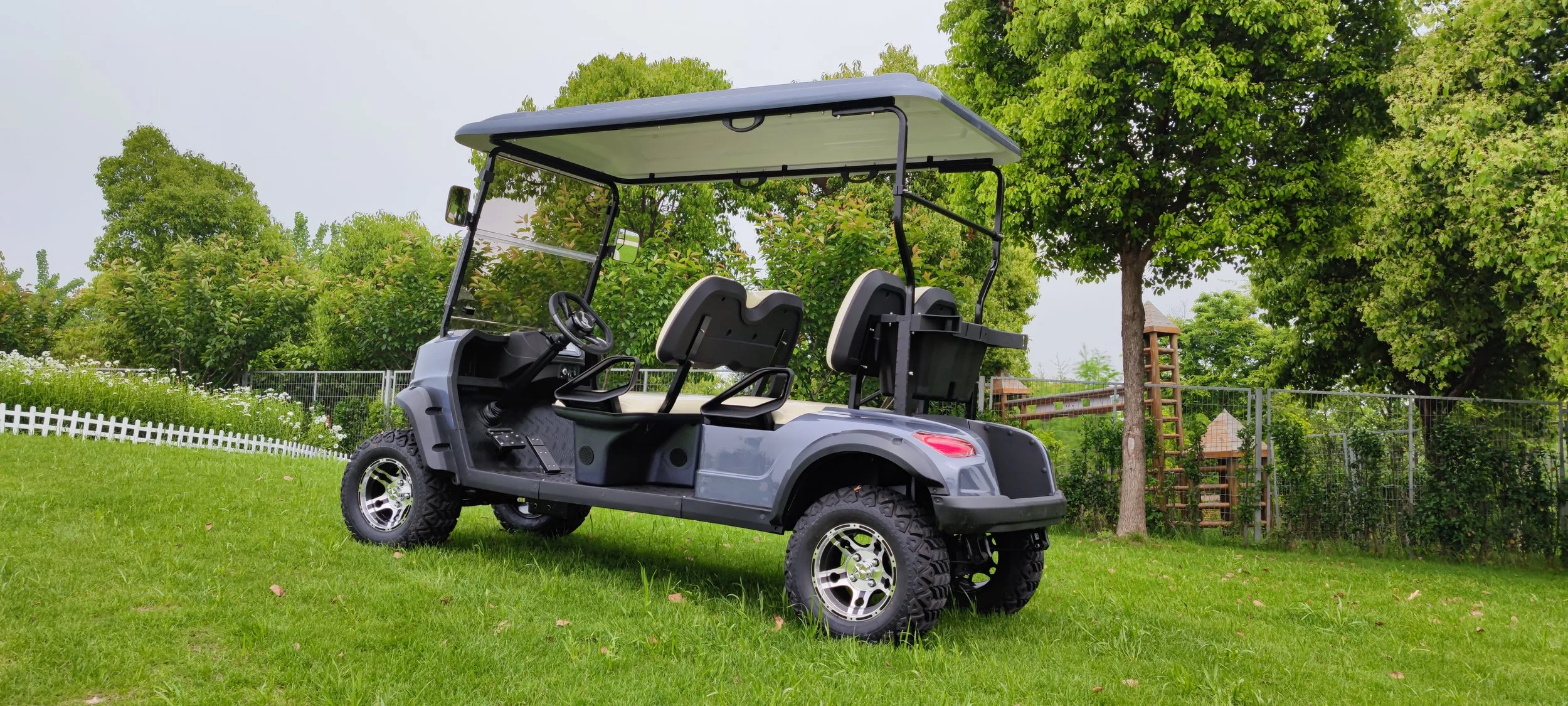 Gray golf cart with four seats and a roof, parked on a grassy area with trees and a fence in the background.
