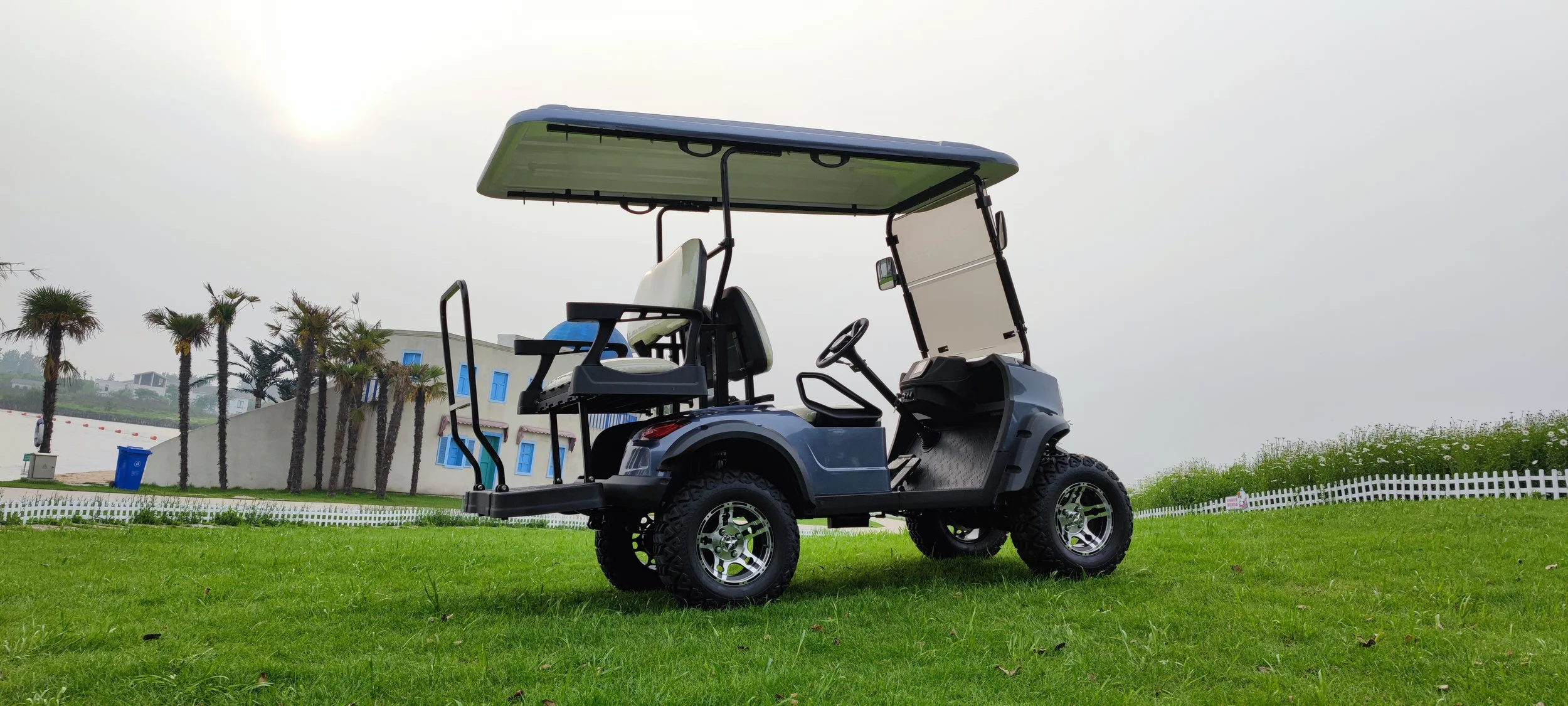 Gray golf cart with white seats parked on green grass, with trees and white fence in the background, under a cloudy sky.
