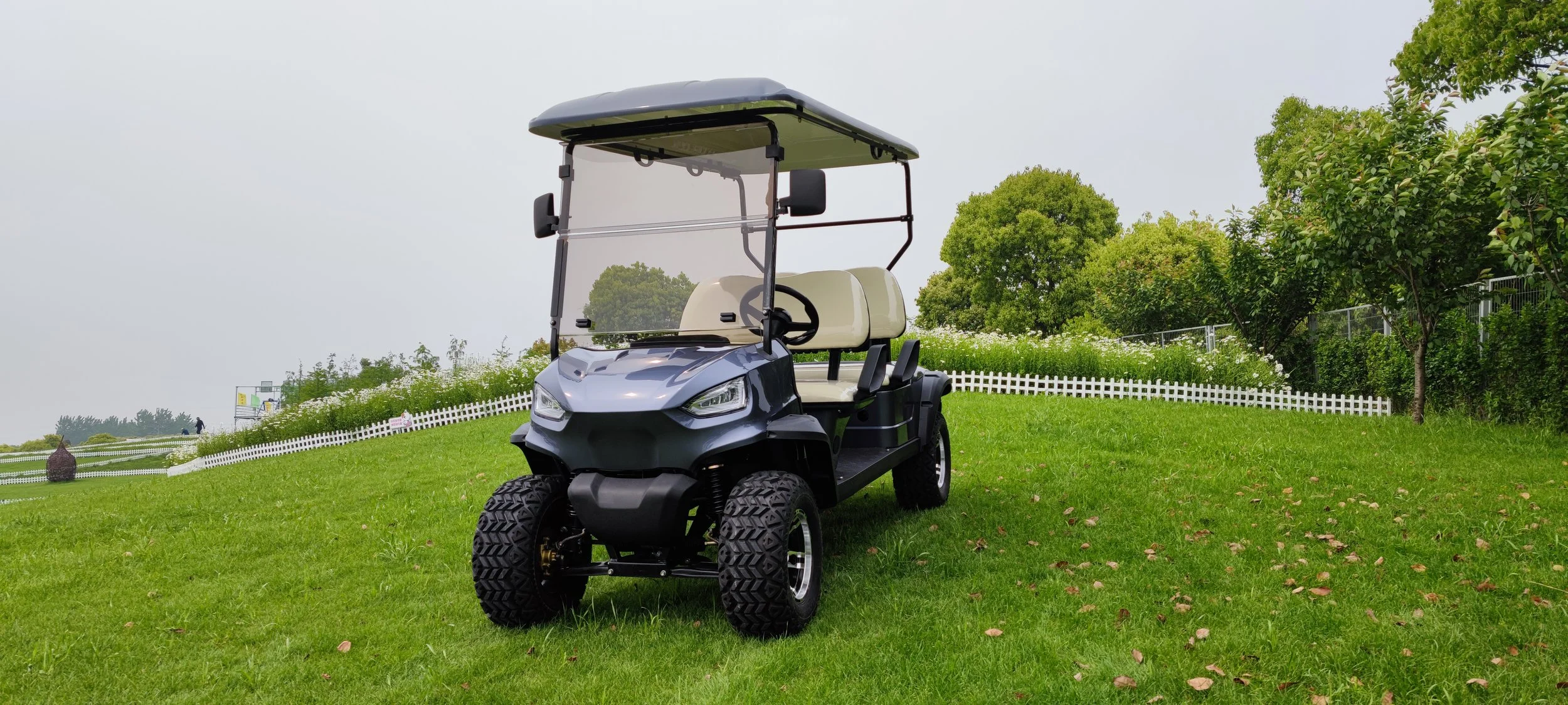 A silver golf cart parked on a grassy field with trees and white picket fences in the background.