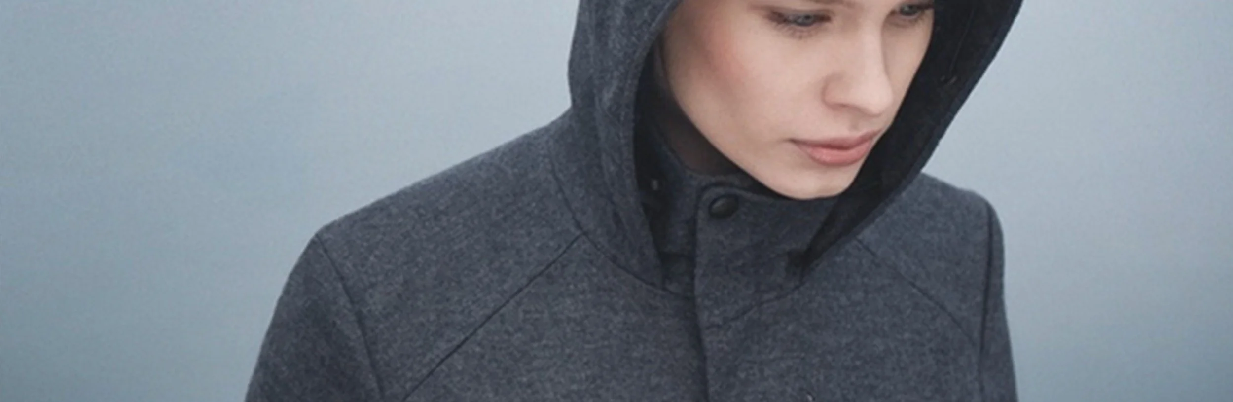 Close-up of a woman wearing a gray hooded jacket, looking down with a neutral expression.