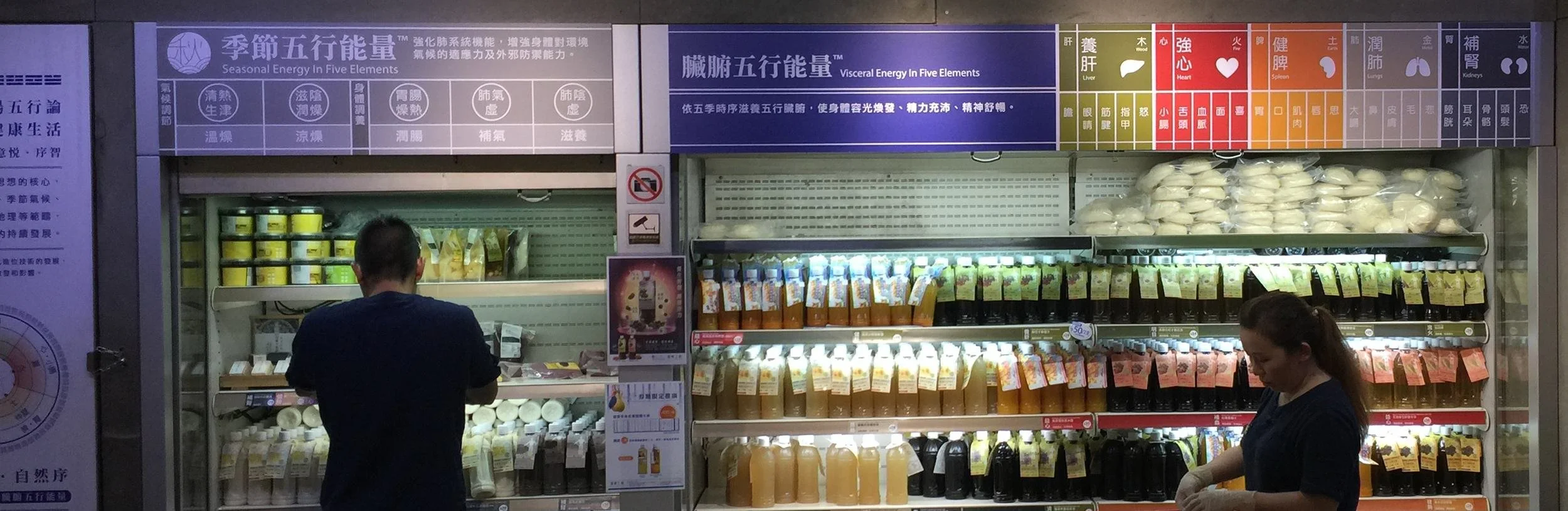 A refrigerated section in a store with various bottled and packaged drinks and food items, with two people shopping in front of it. There's signage in Chinese characters and a colorful informational sign on top.
