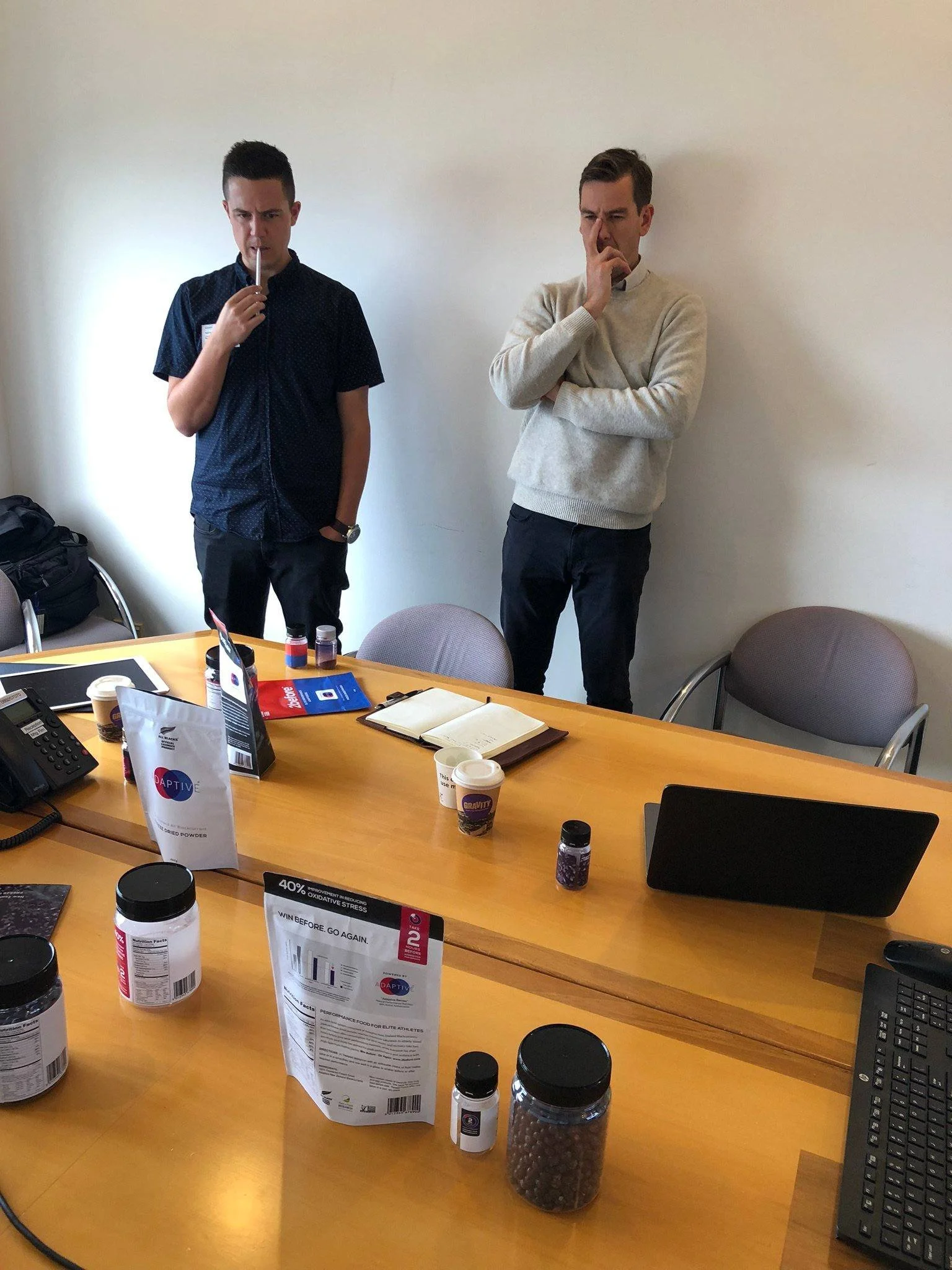 Two men standing in a conference room, one holding a thermometer to his nose and the other with his finger on his nose, surrounded by tables with health supplement bottles, pamphlets, notebooks, and a laptop.