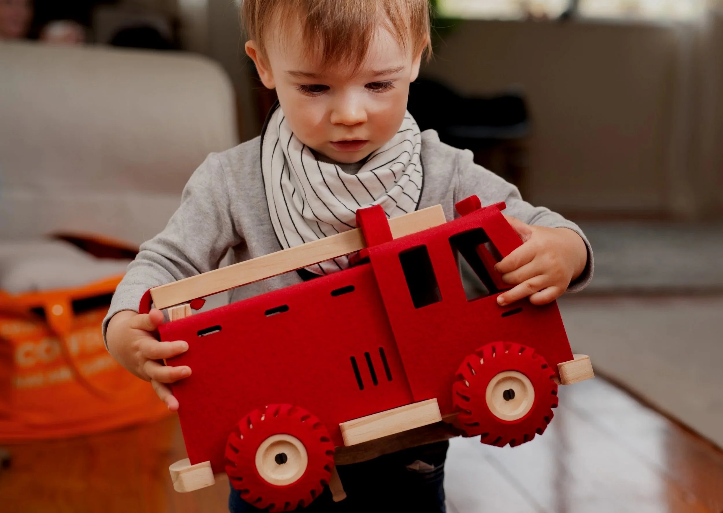 A young boy with red hair and a striped scarf playing with a red and wooden woolkin toy fire truck indoors.