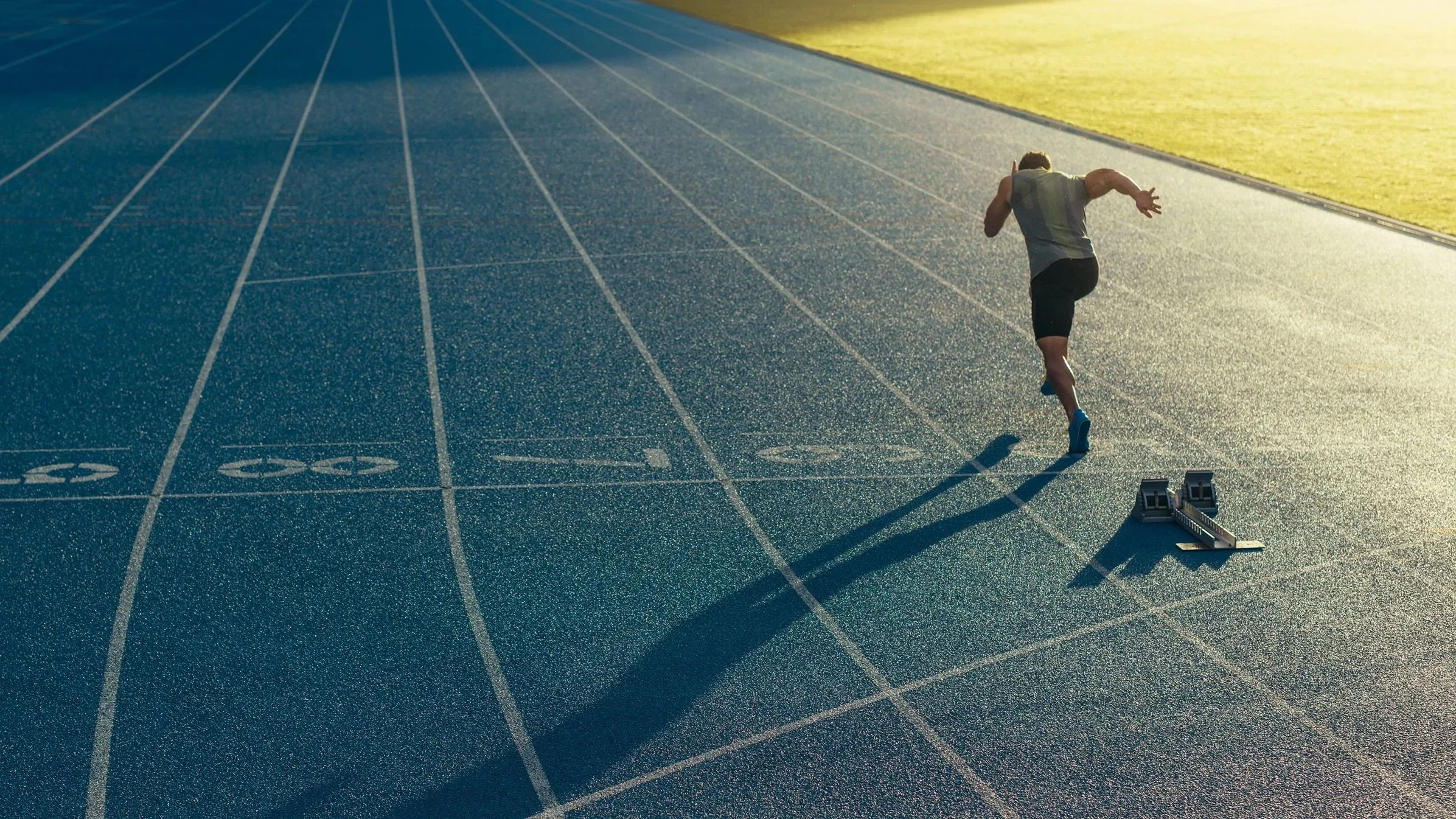 A male athlete running on a blue outdoor track with numbered lanes, some equipment nearby, and the sun casting long shadows during sunset.