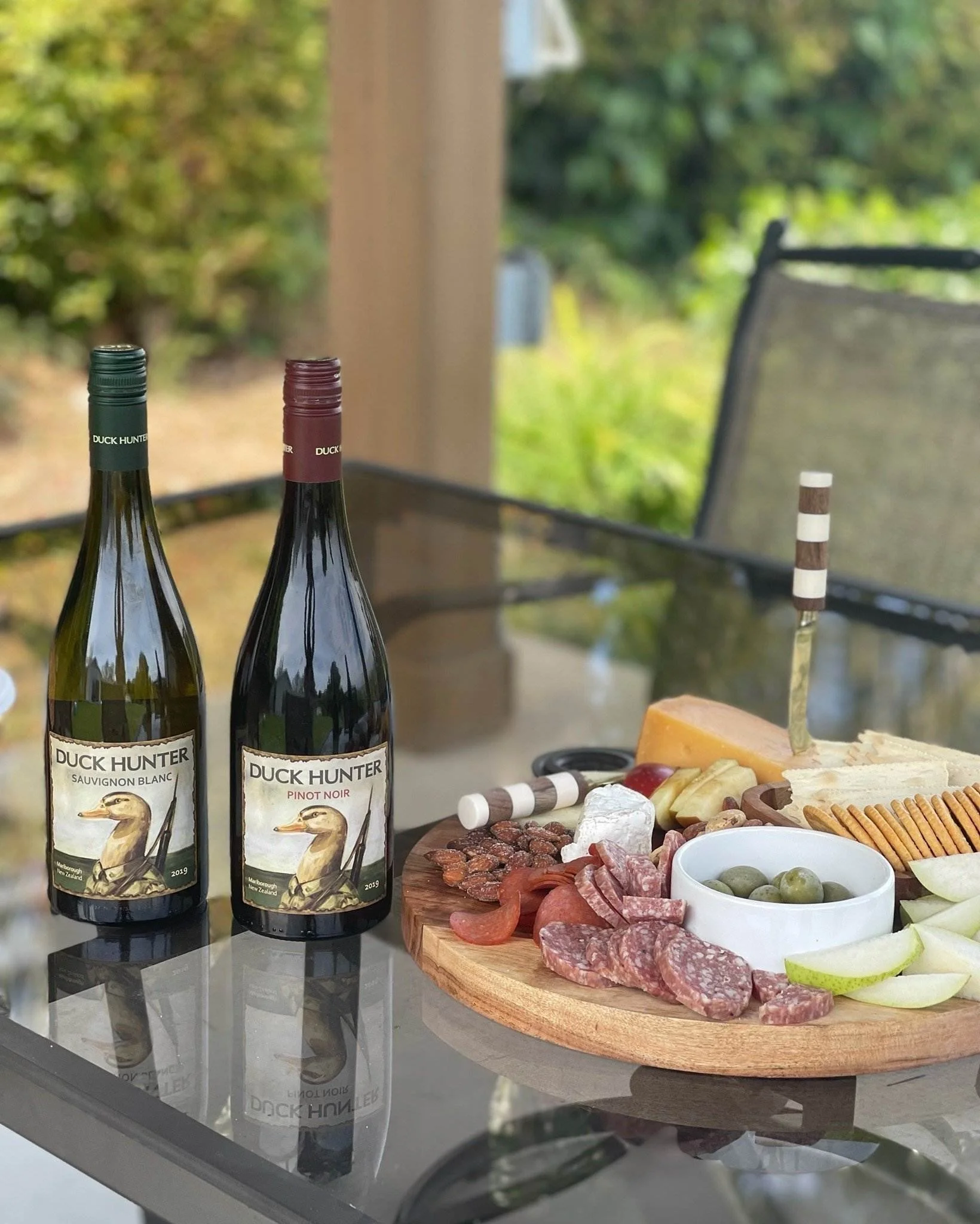 Three bottles of Duck Hunter wine on a reflective table, a charcuterie board with sliced meats, cheeses, and crackers, and a bowl of green olives and lime wedges, with outdoor greenery in the background.