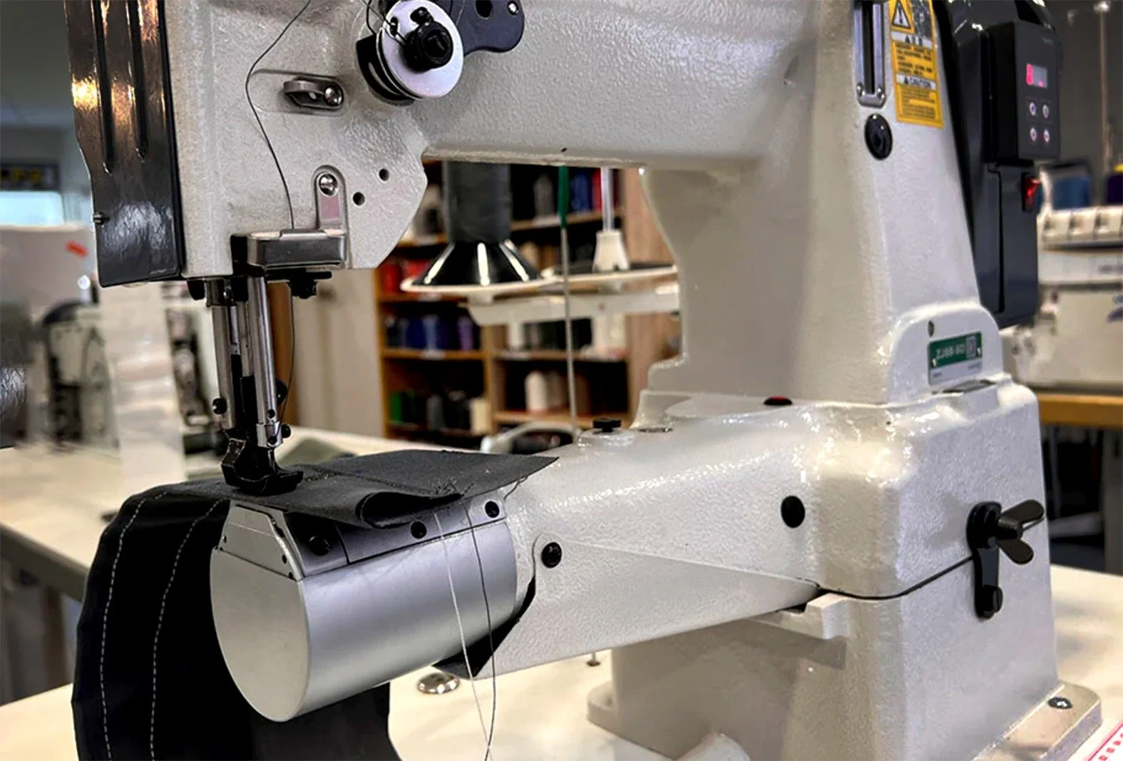 Close-up of a industrial sewing machine in a workshop, with a piece of black fabric under the needle, and a background of shelves with various items.