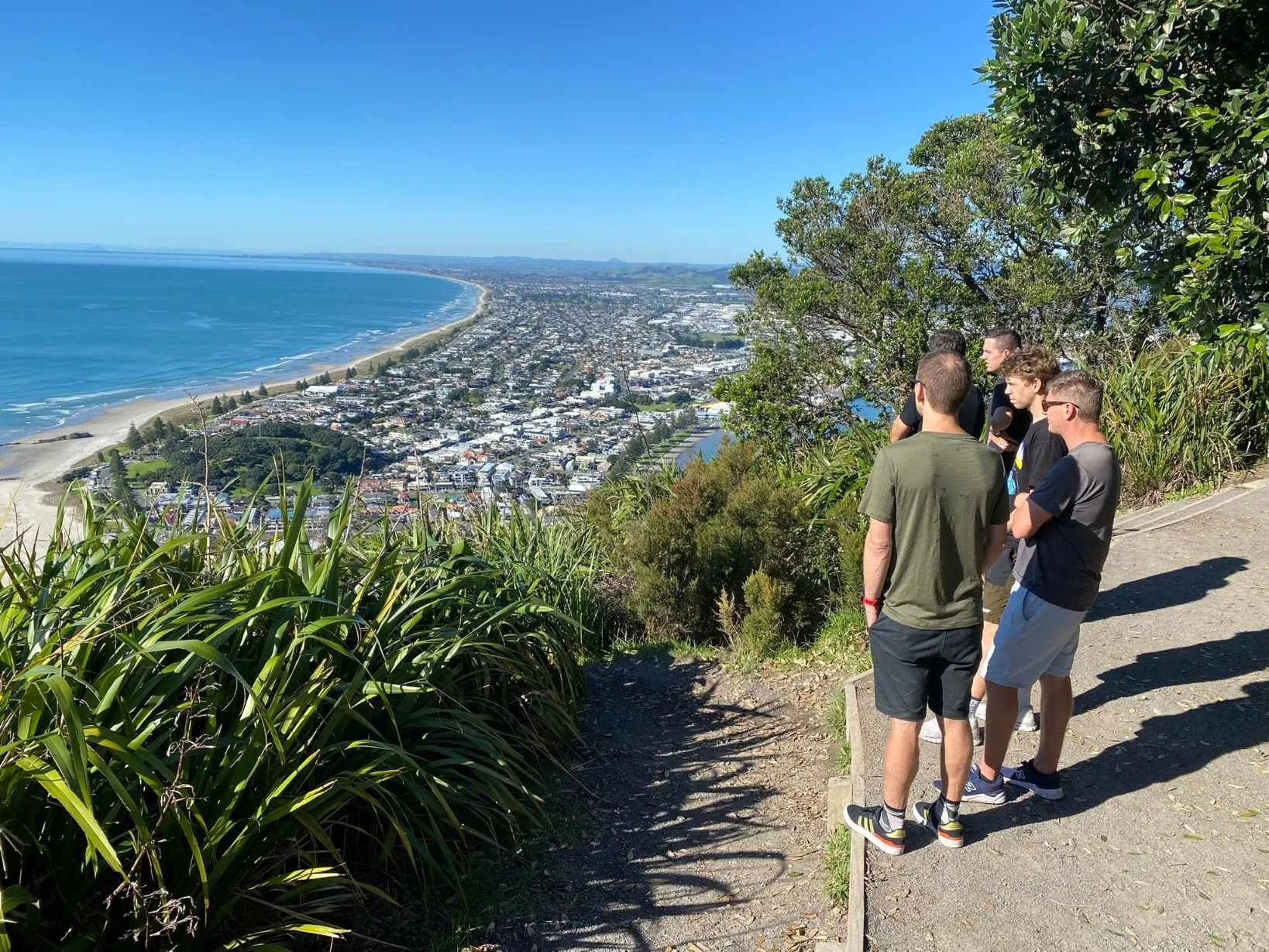 Group of young men standing on a hiking trail overlooking a coastal town, beach, and ocean under a clear blue sky.