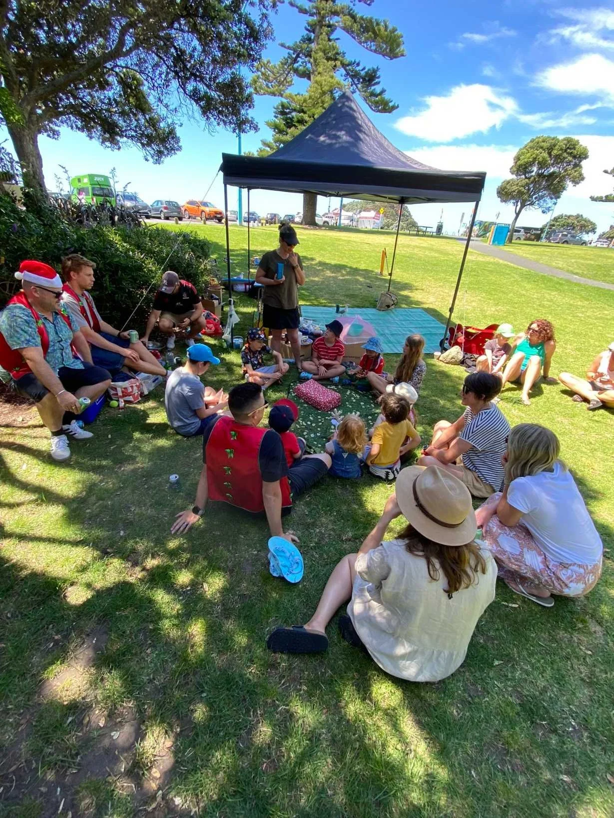 People gathered outdoors under a canopy in a park, sitting on the grass, some wearing hats and some in holiday attire, around a person who appears to be leading an activity or discussion.