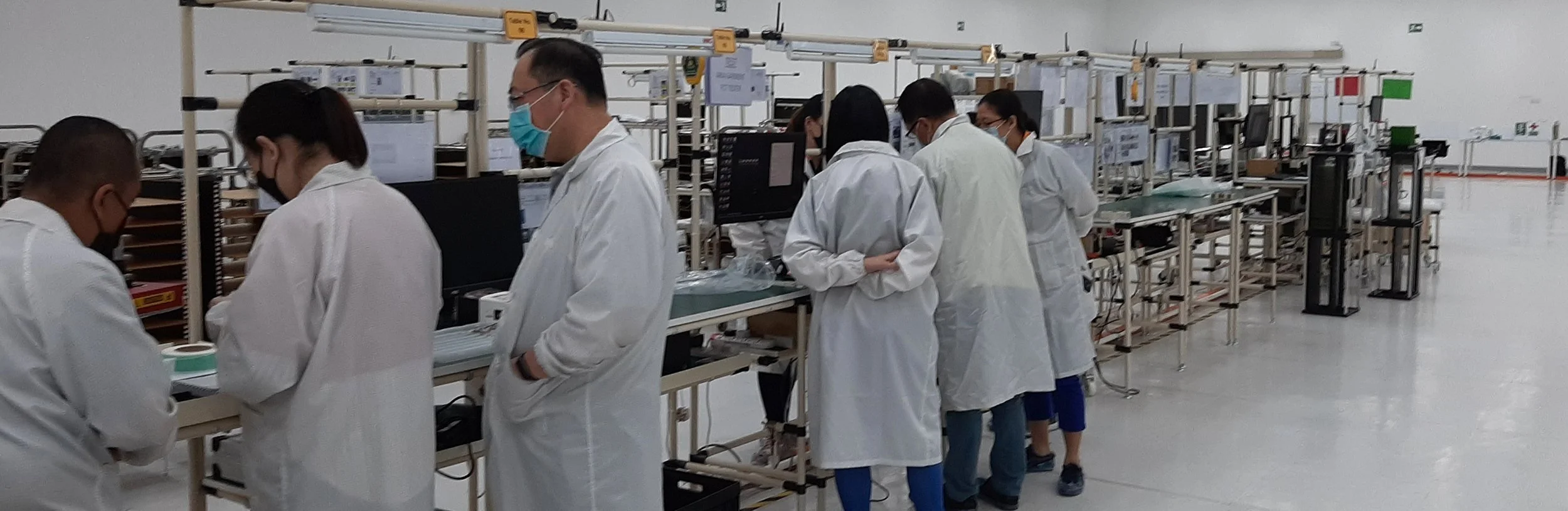 A group of scientists working in a cleanroom assembly line, wearing white lab coats and face masks, surrounded by electronic equipment and components.