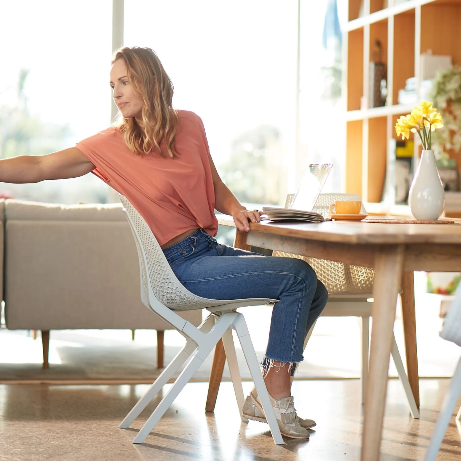 A woman with shoulder-length wavy hair sitting at a wooden dining table in a bright, sunlit room, reaching out with her left arm. There are books, an open laptop, a yellow flower vase, and a coffee cup on the table.