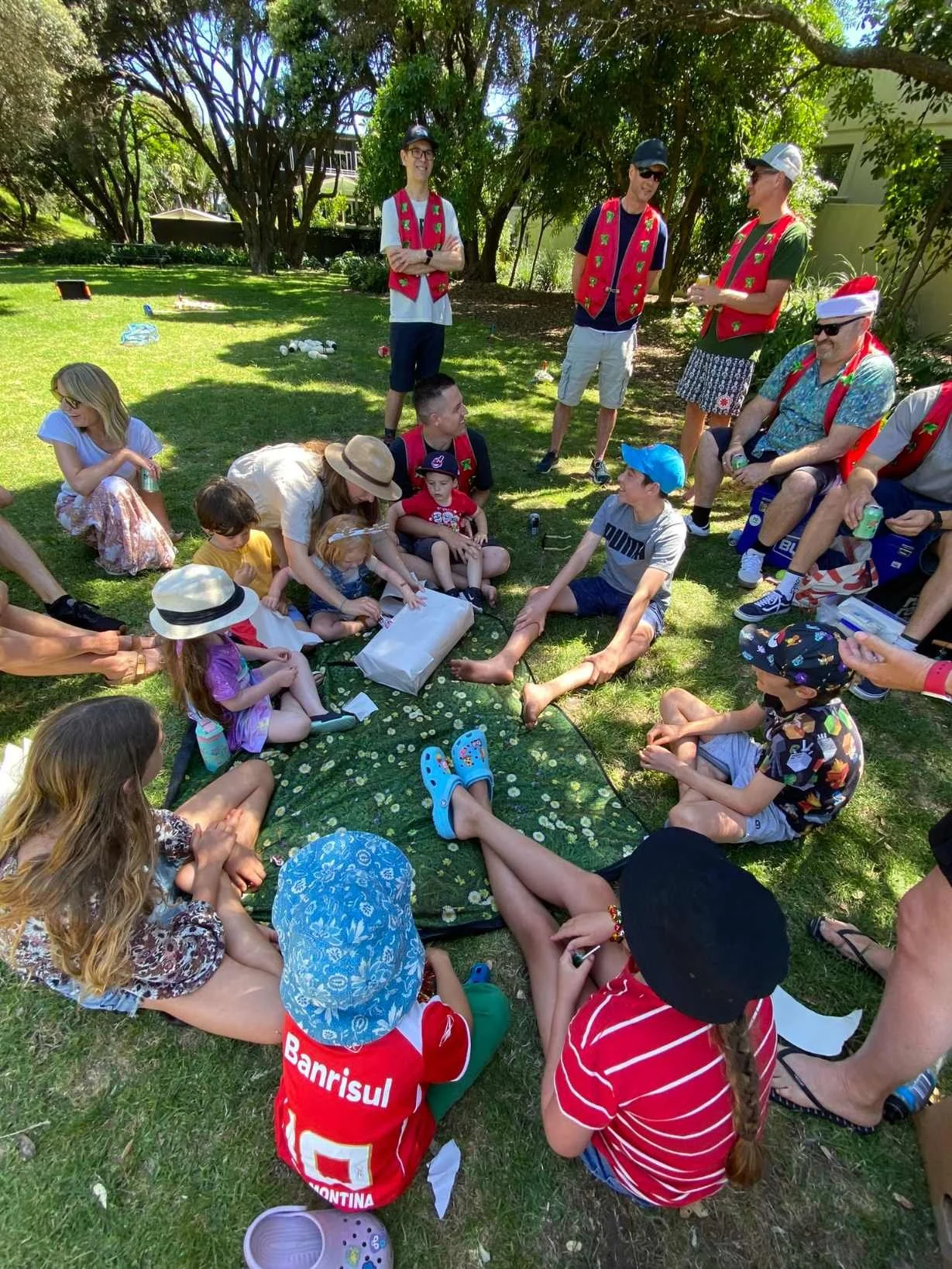 Group of children and adults sitting on the grass outdoors, gathered around a woman passing out gifts, with others standing and sitting nearby, some wearing festive clothing and hats, in a sunny park with trees and a building in the background.