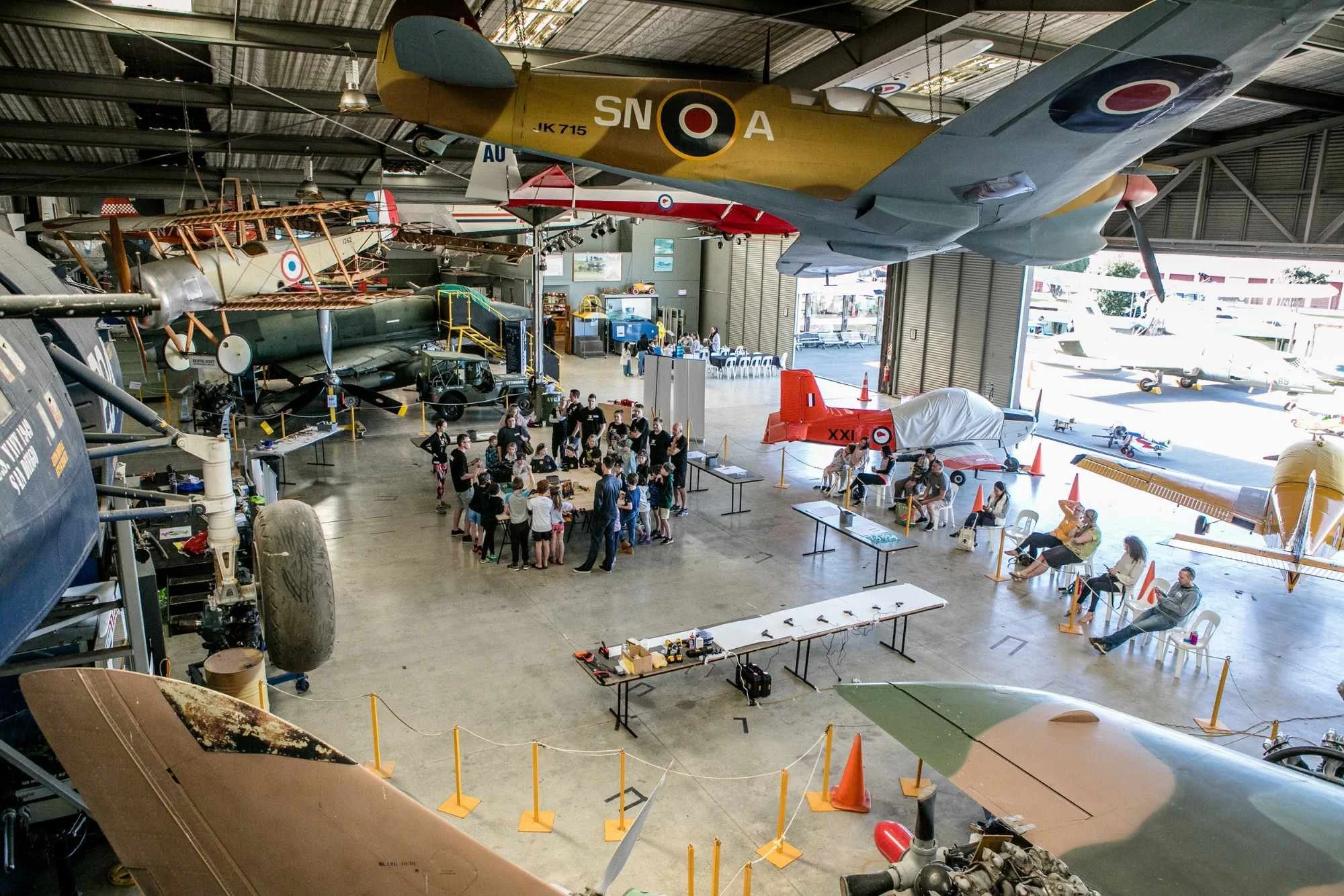 Museum hangar with vintage aircrafts and exhibits. Visitors walk and sit at tables, with some aircraft parts and models displayed.