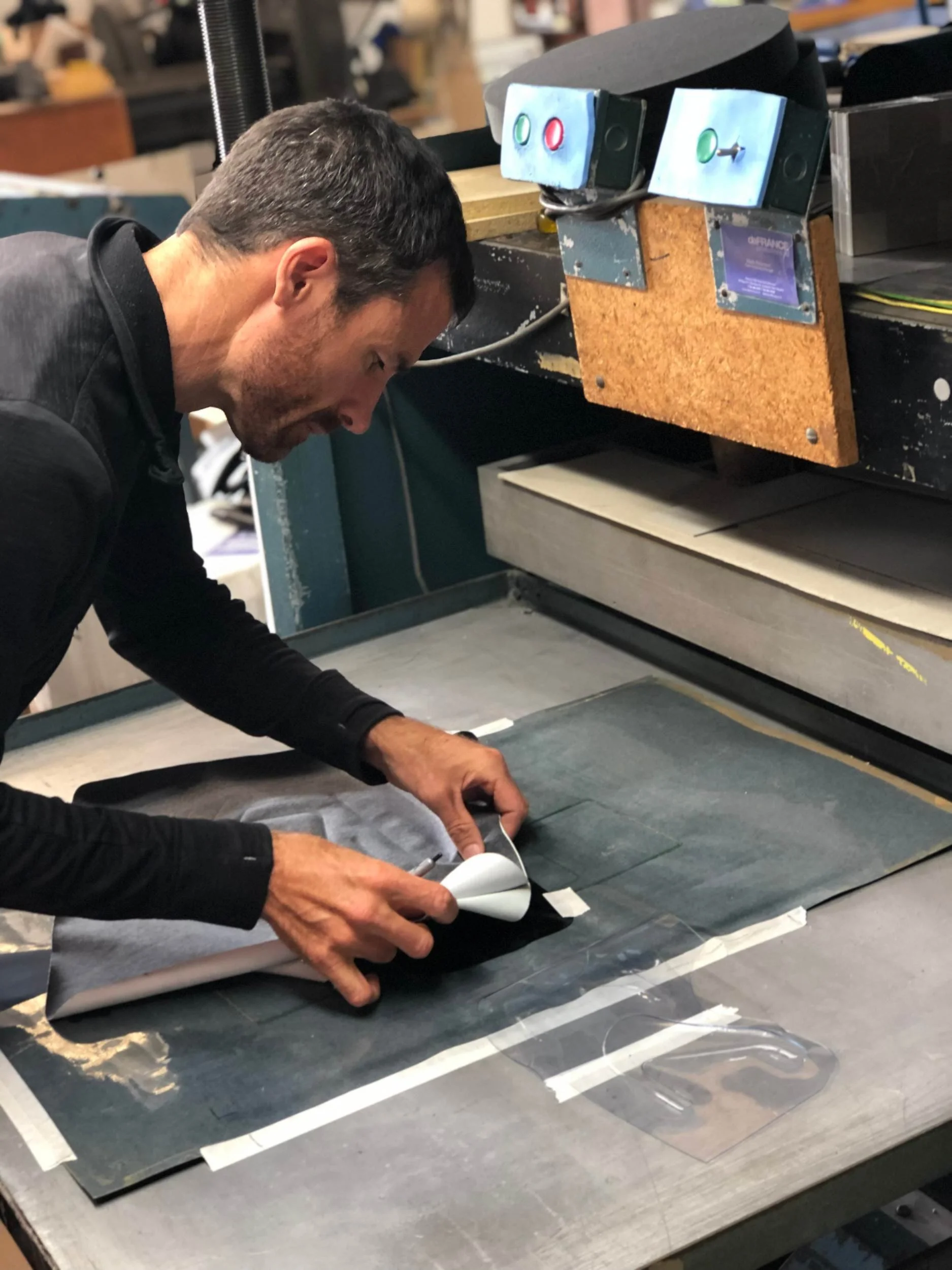 A man is working on a project at a workbench in a workshop, using a paper template or pattern on a sheet of dark material, with various tools and equipment around him.