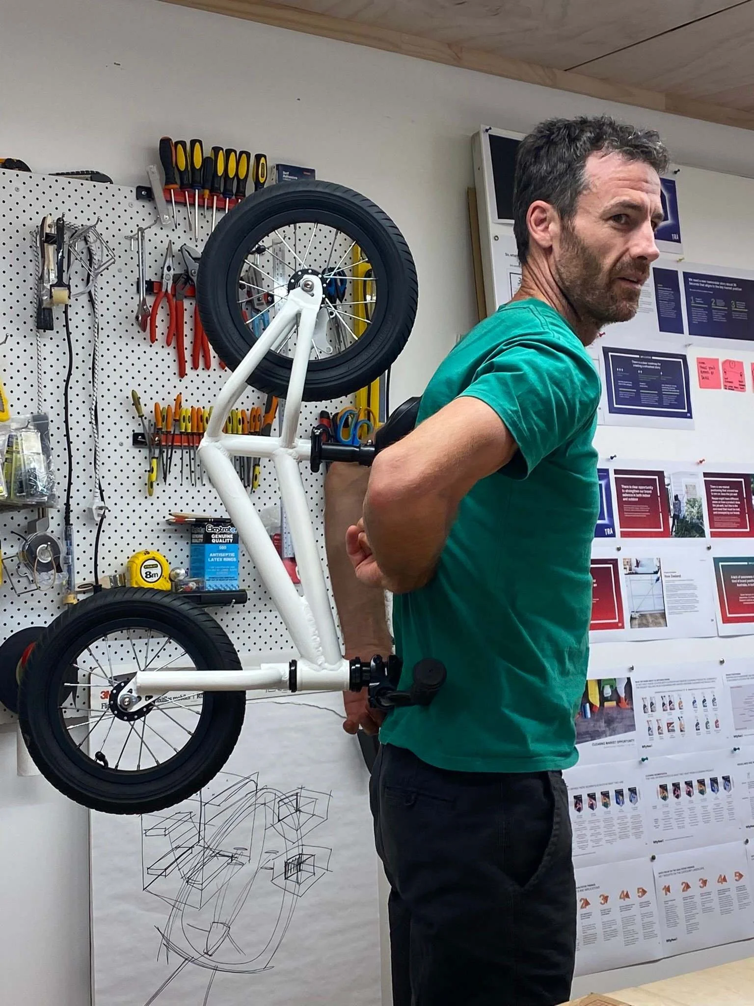 A man in a green shirt stands in front of a pegboard with various hand tools and a partially assembled white bicycle. The workspace has posters and diagrams on the wall.