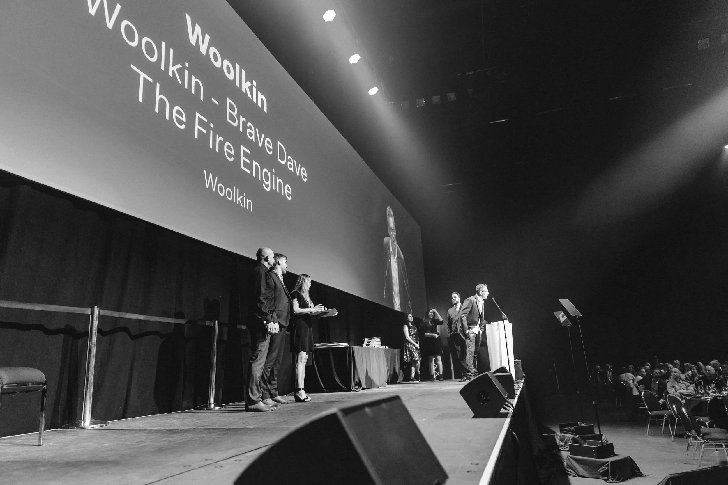 A group of people standing on stage at an event, with a large screen behind them displaying text about Woolkin, Bravex Dave, The Fire Engine, and Woolkin. Audience members are seated at tables in the foreground.