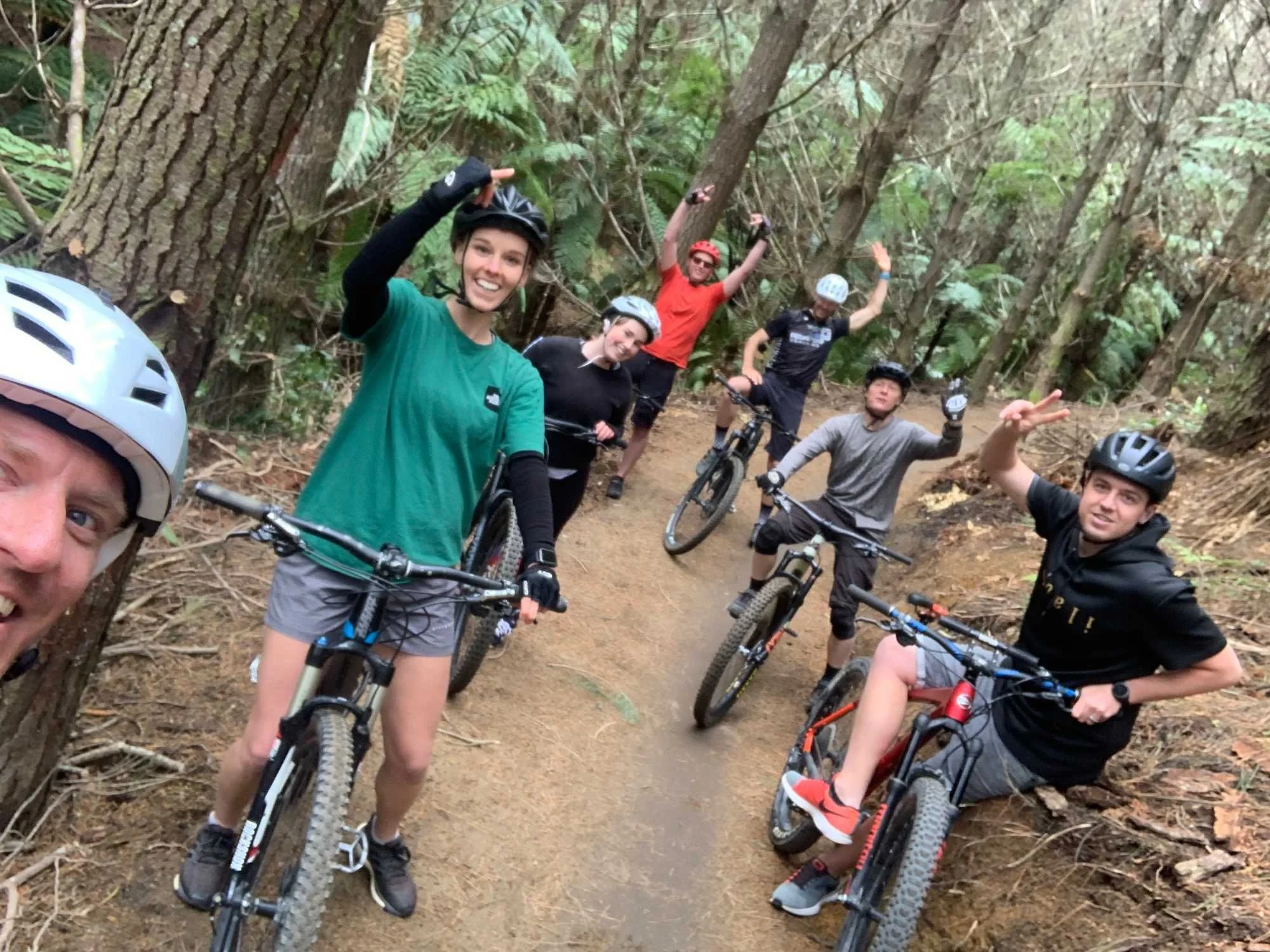 Group of six people on mountain bikes in a forest, wearing helmets and casual outdoor clothing, smiling and posing for a photo on a dirt trail surrounded by trees and greenery.