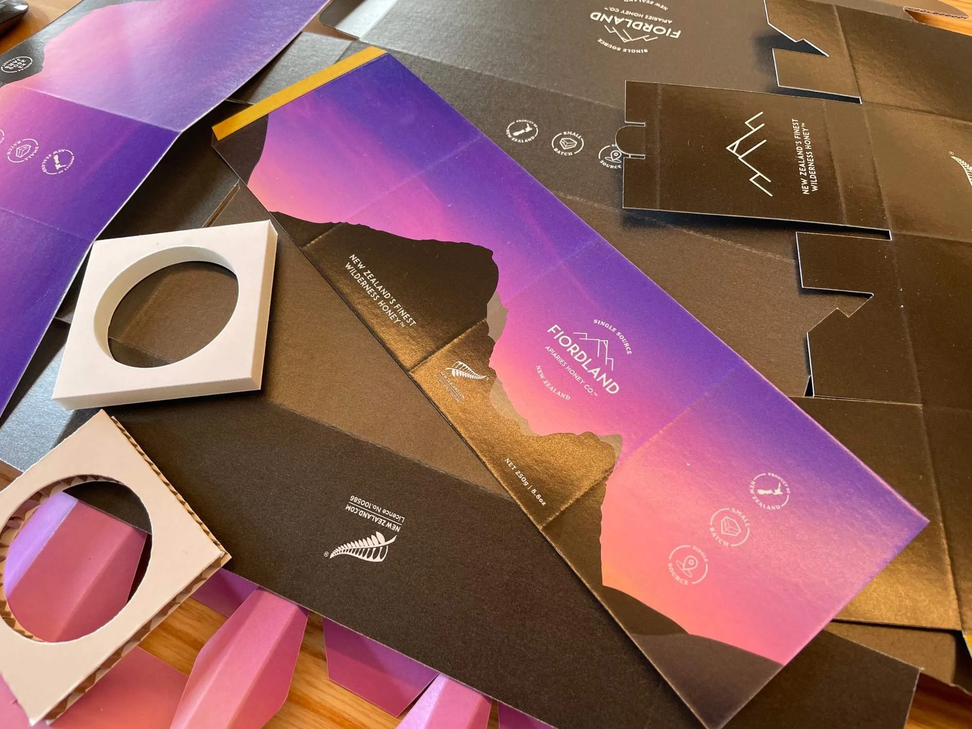 Various cardboard boxes with purple and black exterior, featuring New Zealand wilderness honey branding and icons indicating sustainability and source, some with pink handles.