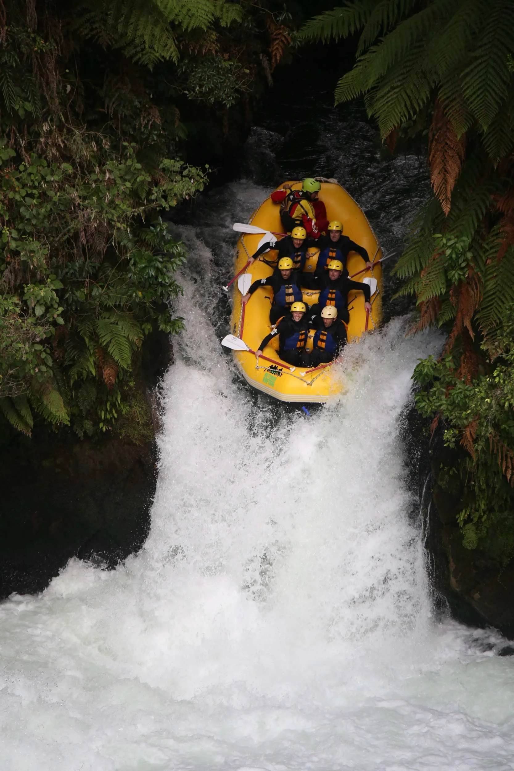 Group of people wearing helmets and safety gear white rafting on a river surrounded by lush green foliage.
