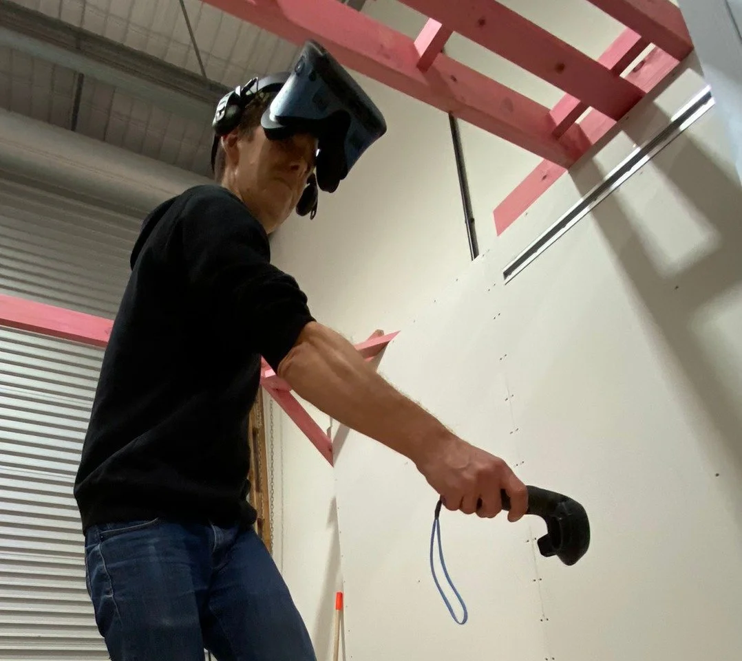 A man wearing a black shirt and blue jeans is working on construction, standing next to a white wall, using a power tool. He is wearing a headset and a virtual reality headset. Pink construction materials are attached to the ceiling, and a ladder is visible on the right side of the image.