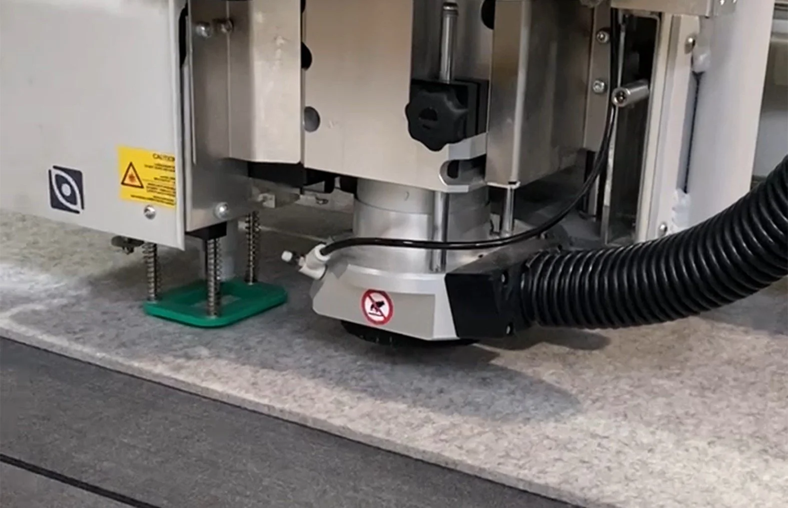 Close-up of machinery showing metallic components, a warning label, an optical sensor, a black cable, and a flexible black hose, on a gray surface.