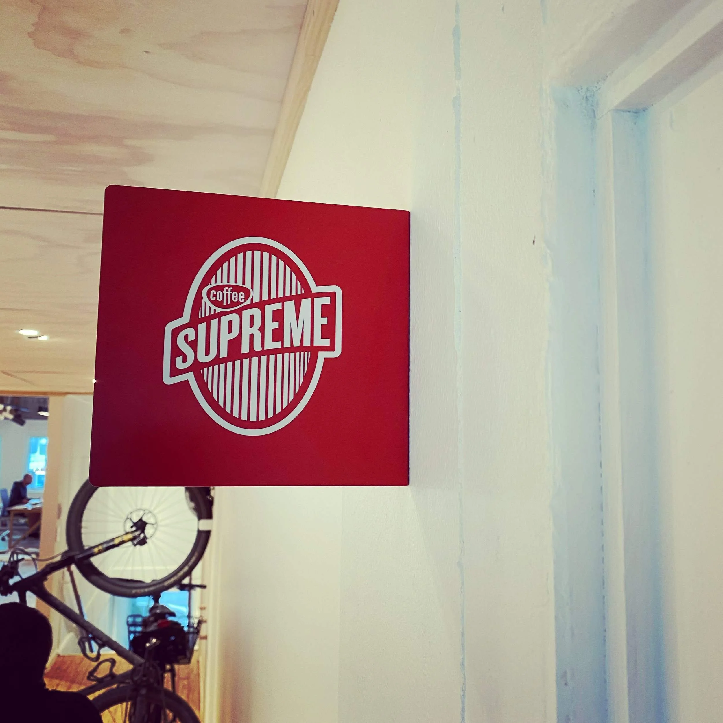 A red sign with the Supreme logo in white, part of a bicycle is visible in the foreground, and the background shows a light yellow wall and a wooden ceiling with lights.