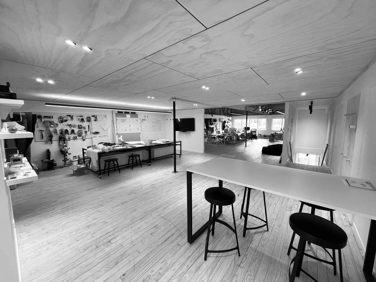 A spacious modern office or workshop with wooden flooring and ceiling. It features workstations, a wall with tools and supplies, a large whiteboard, a mounted TV, and a mix of seating including bar stools and chairs. There are large windows in the background letting in natural light.