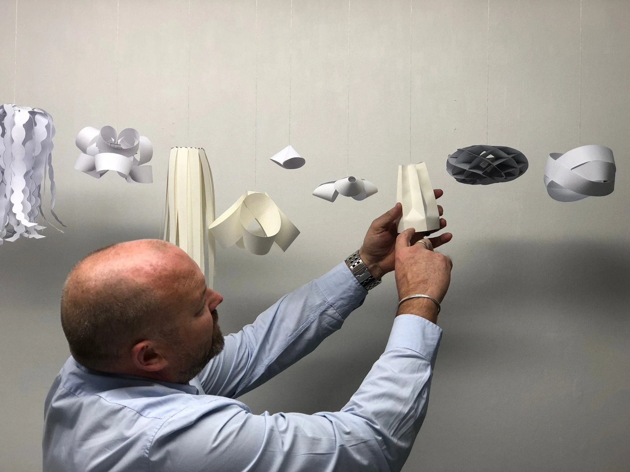 A bald man with a beard and wearing a light blue shirt is holding a white geometric paper lamp. Other paper lamps with different shapes and colors are hanging against a plain wall.