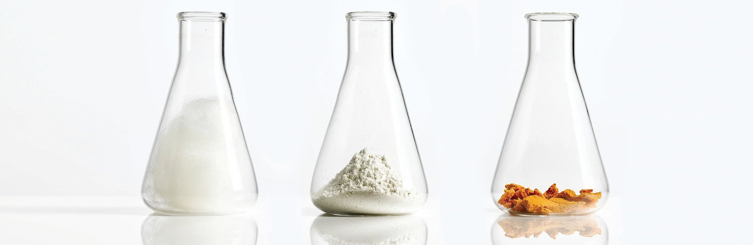 Three glass beakers with powders and spices inside, from left to right: a white powder, a light-colored powder, and orange spices on a white background.