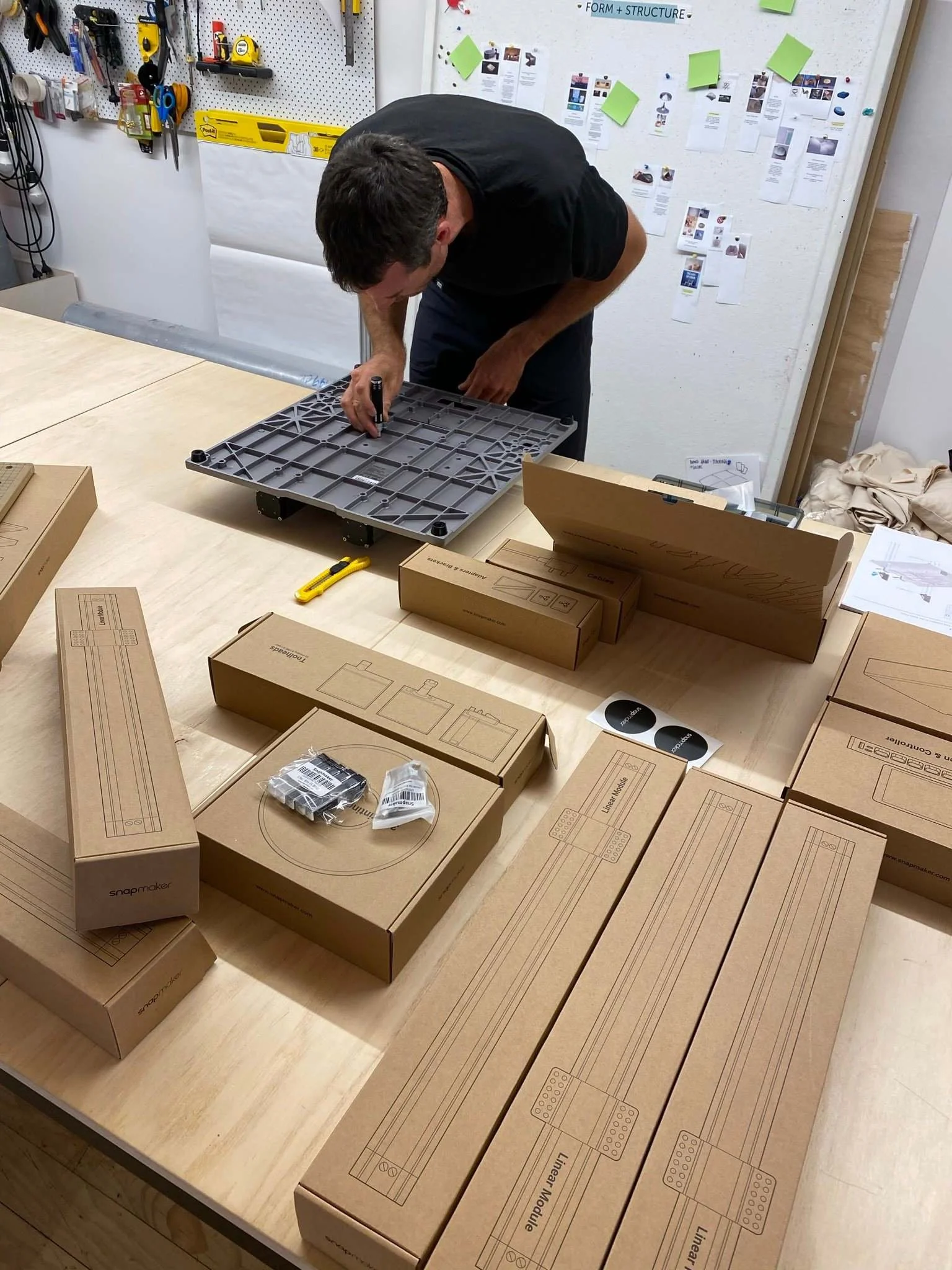 A person working on assembling a piece of furniture in a workshop, surrounded by cardboard boxes labeled 'snapmaker' and various tools on the pegboard wall.