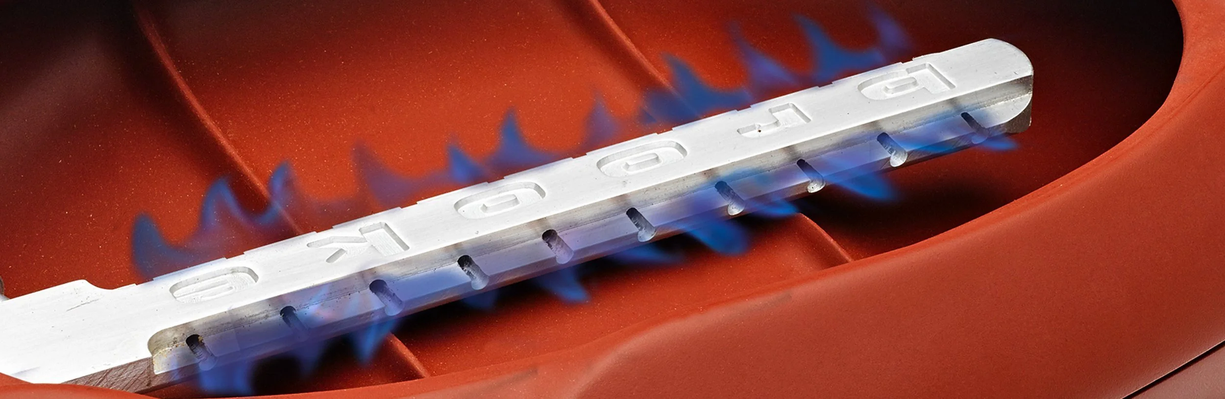 Close-up of a metal component with cut-out letters, mounted on a red surface, with blue flames from a gas torch underneath.