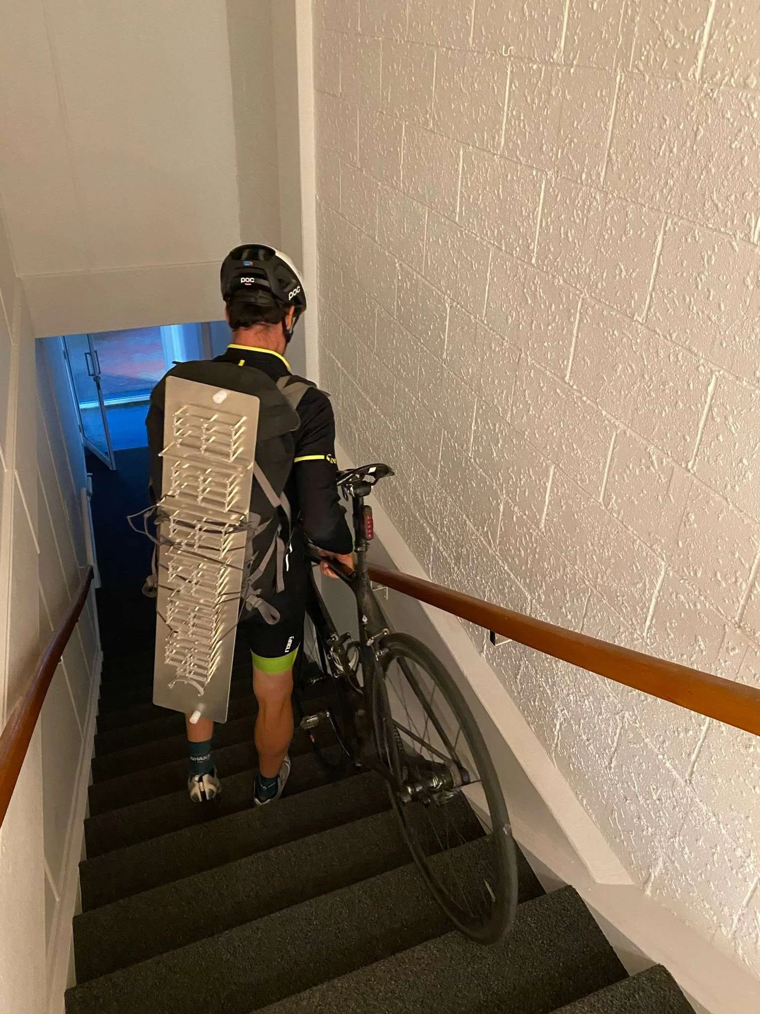 A cyclist wearing a helmet and black cycling gear is walking down a staircase with a mountain bike. He has a large white backpack with ventilation slots. The staircase has black steps and a wooden handrail on the right side. The wall on the right is 