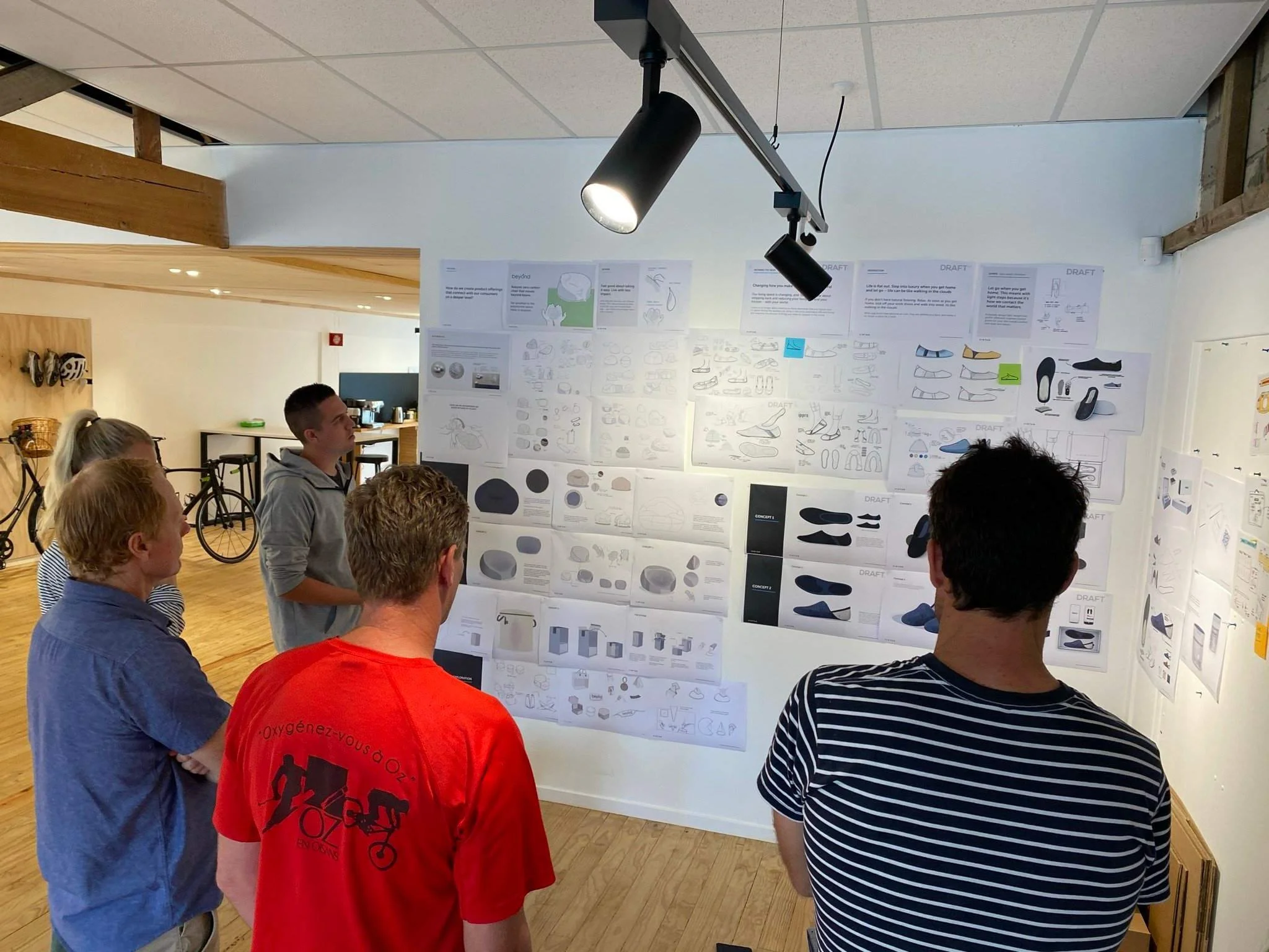 Group of people standing in front of a wall covered with sketches and diagrams, discussing project designs in a room with wooden ceiling beams and ceiling lights.