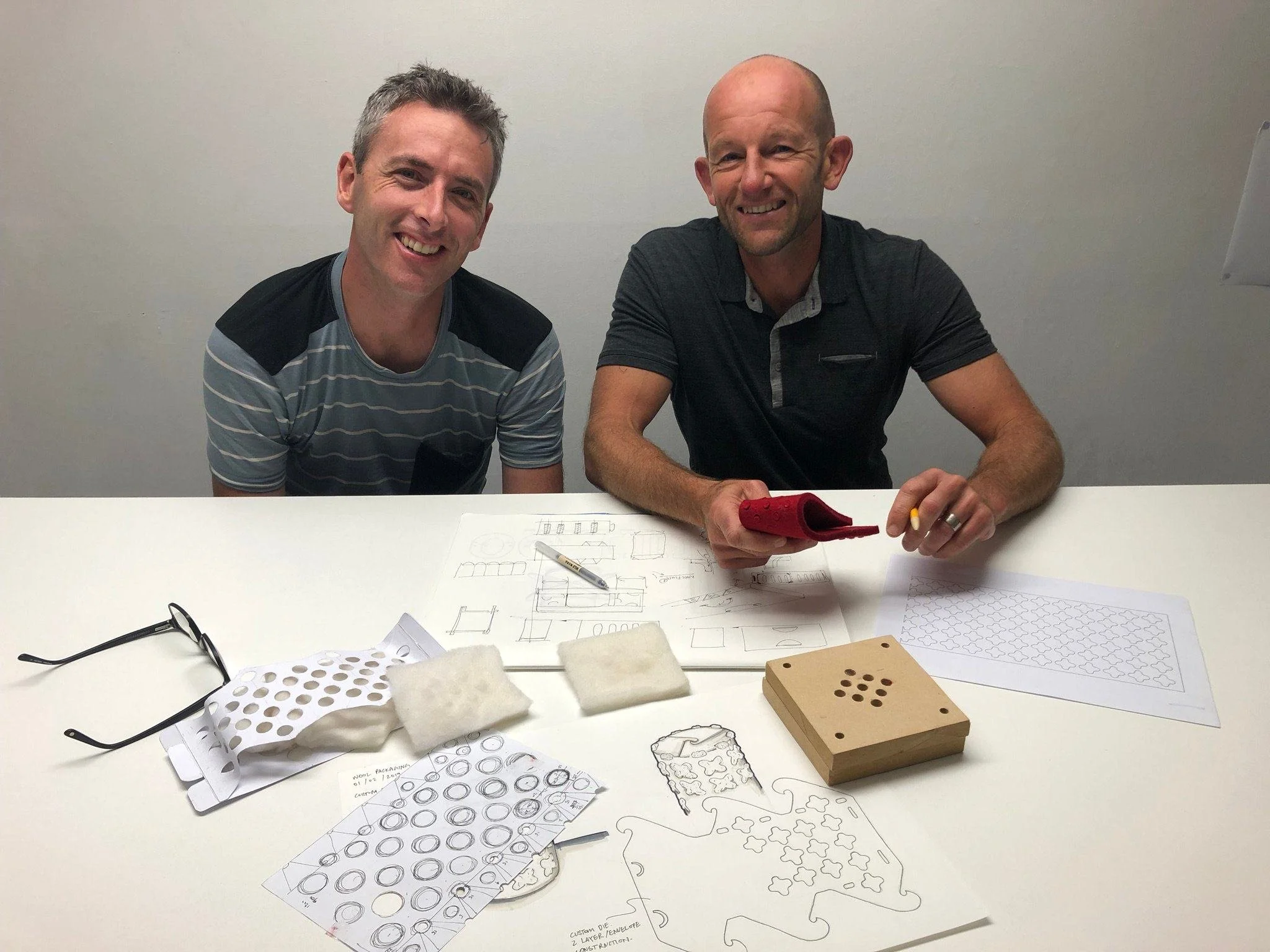 Two men are seated at a white table with various sketches, papers, and patterned templates spread out. The man on the left has short, gray hair and is wearing a striped shirt, while the man on the right is bald and has a beard, wearing a dark shirt. 