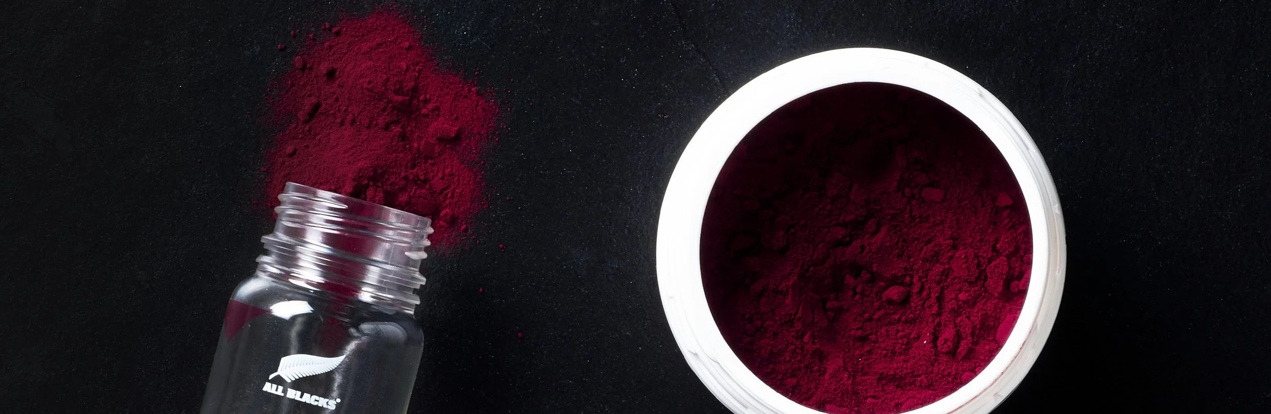 Spilled red powder next to an open container of red powder and a glass jar with a black fern logo.