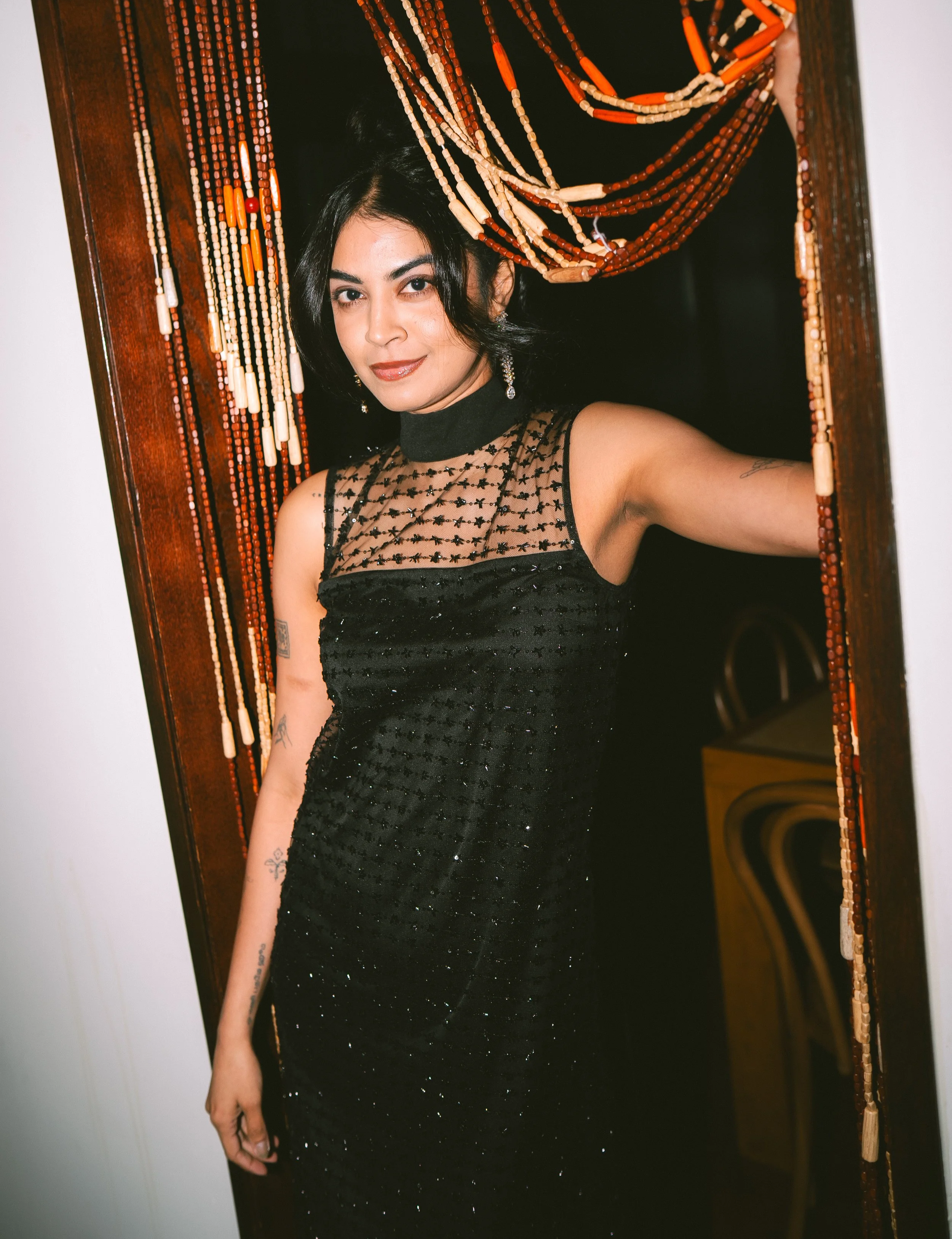 A woman with black hair taking a selfie in a mirror. She is wearing a black, sparkly, semi-sheer dress with a high neckline. She has earrings and tattoos on her arm. The mirror has a beaded frame with shades of brown, orange, and cream beads.