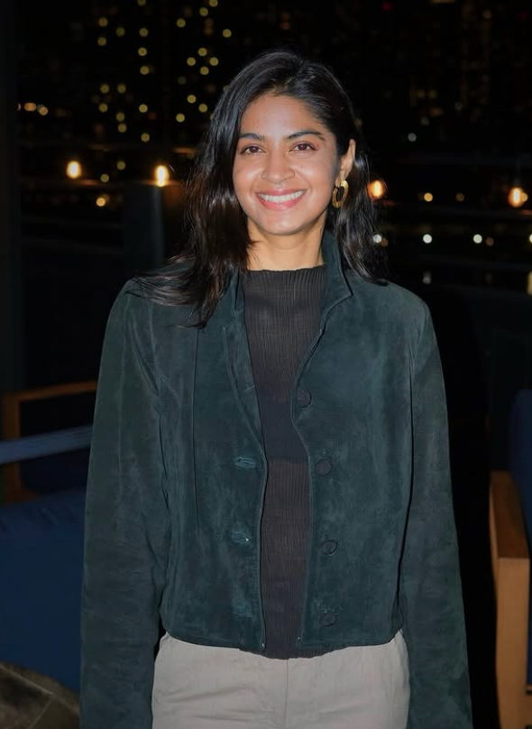 A woman with shoulder-length black hair smiling, wearing a dark green jacket over a black top, standing outdoors at night with blurred city lights in the background.