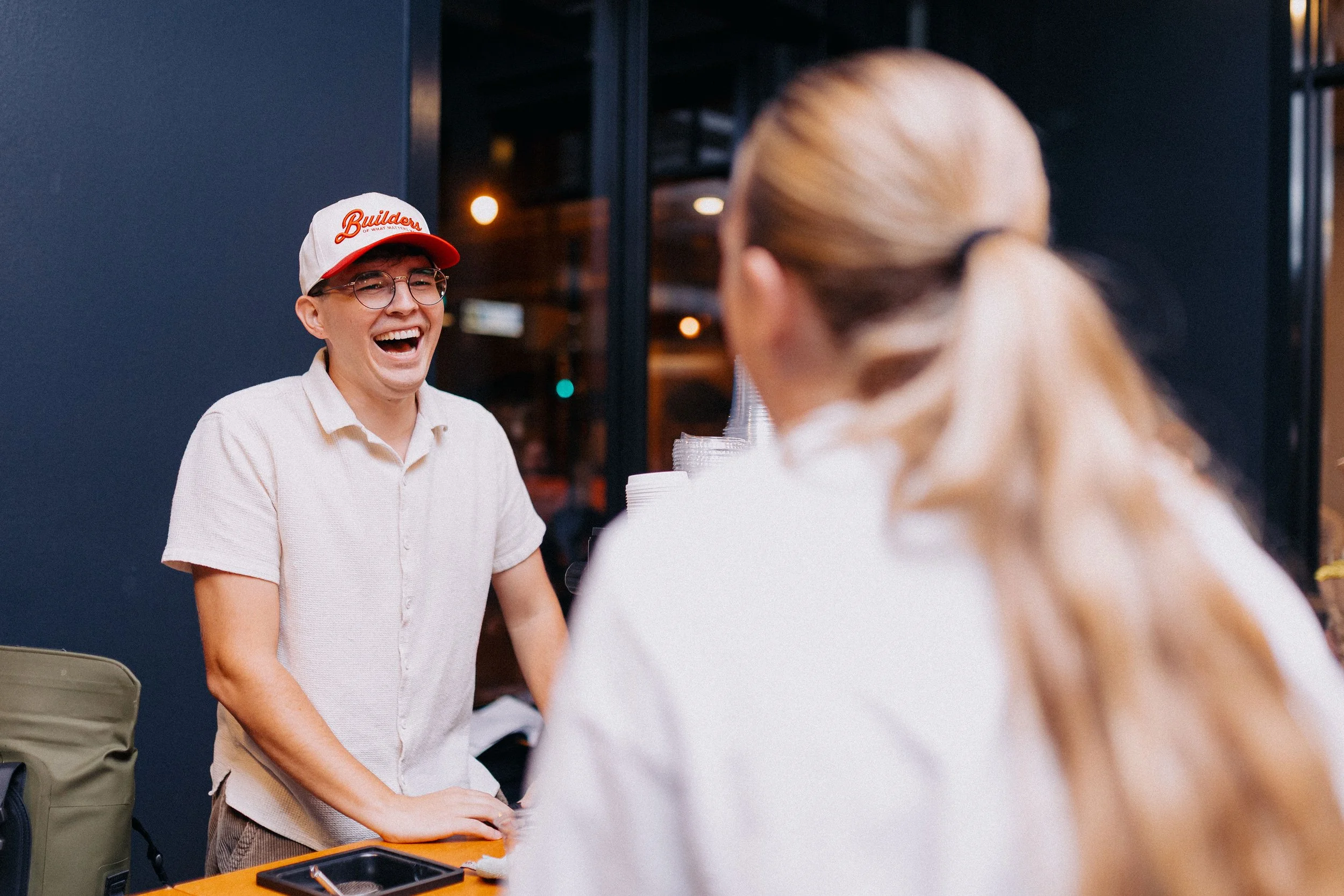 A young man with glasses and a red and white cap laughing and talking to a woman with blonde hair tied back, at an indoor event.