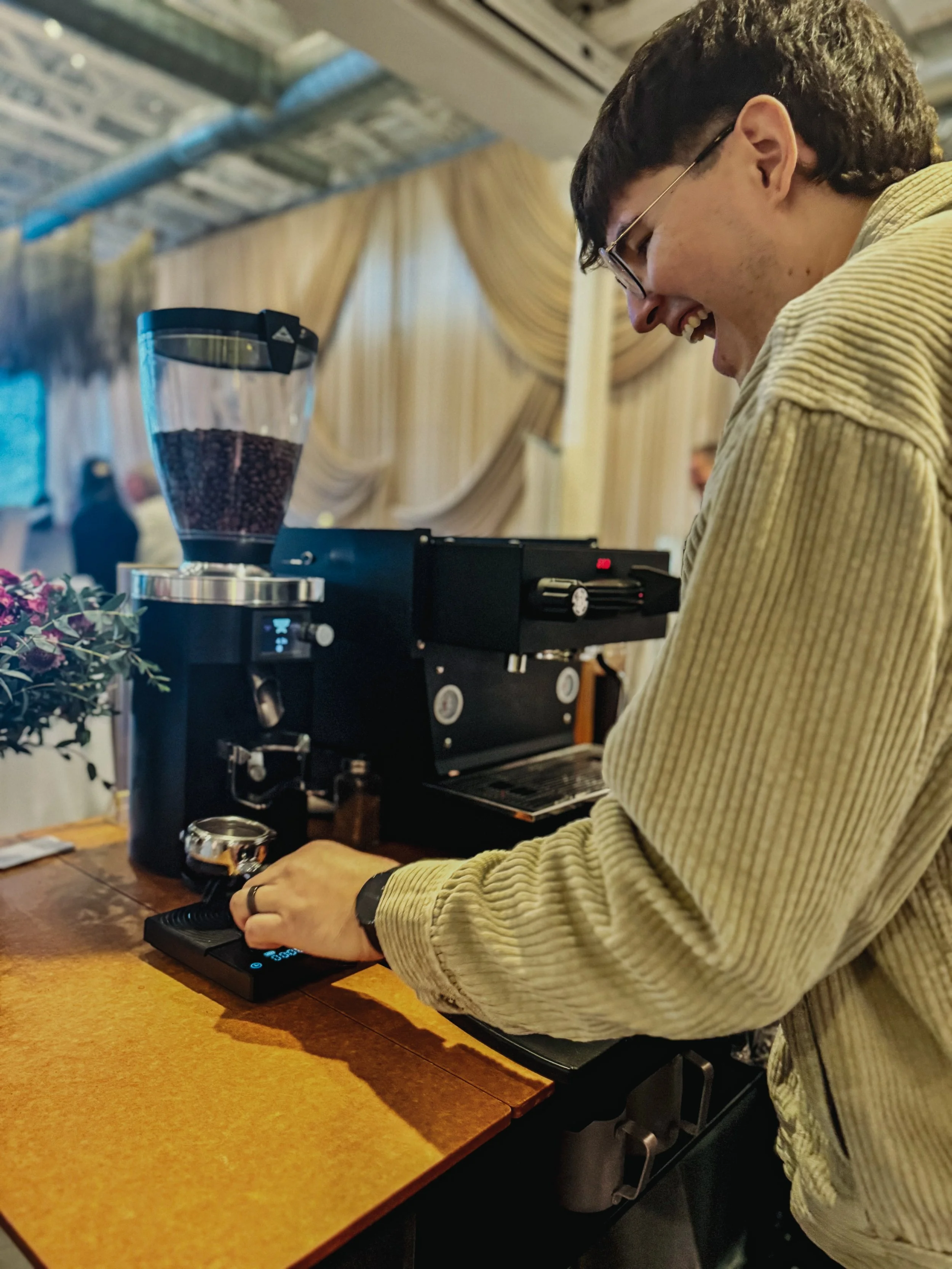 A smiling barista adjusting the espresso machine controls behind a wooden counter with a coffee grinder, a small plant, and a wristwatch.