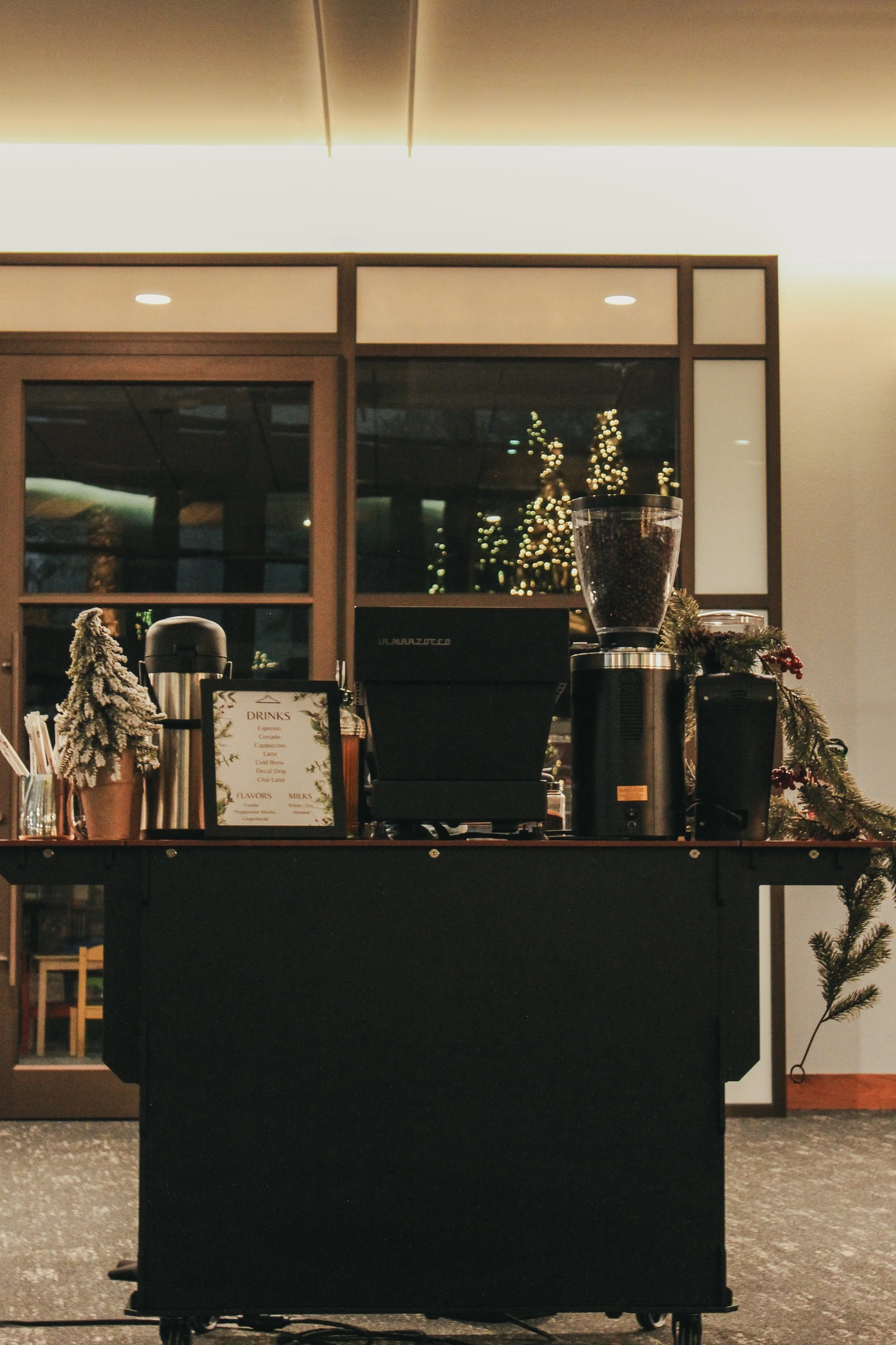 A coffee shop counter decorated with small holiday trees and greenery, with Christmas lights reflecting in the window behind it.